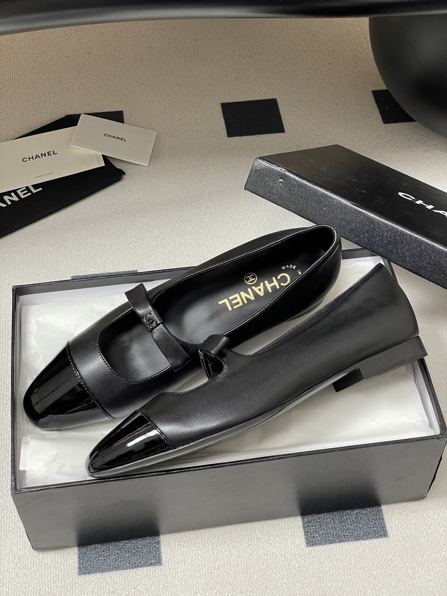 A pair of authentic Chanel black leather Mary Jane flats with glossy patent leather toe caps and CC logo straps displayed on a black surface. - Hình 4