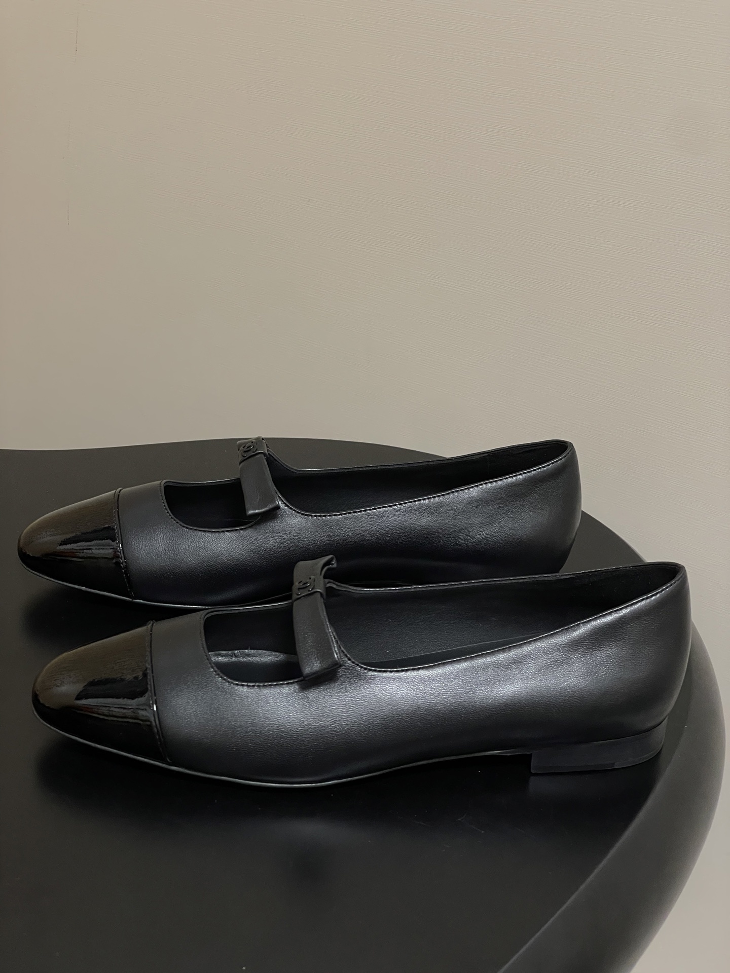 A pair of authentic Chanel black leather Mary Jane flats with glossy patent leather toe caps and CC logo straps displayed on a black surface. - Hình 3