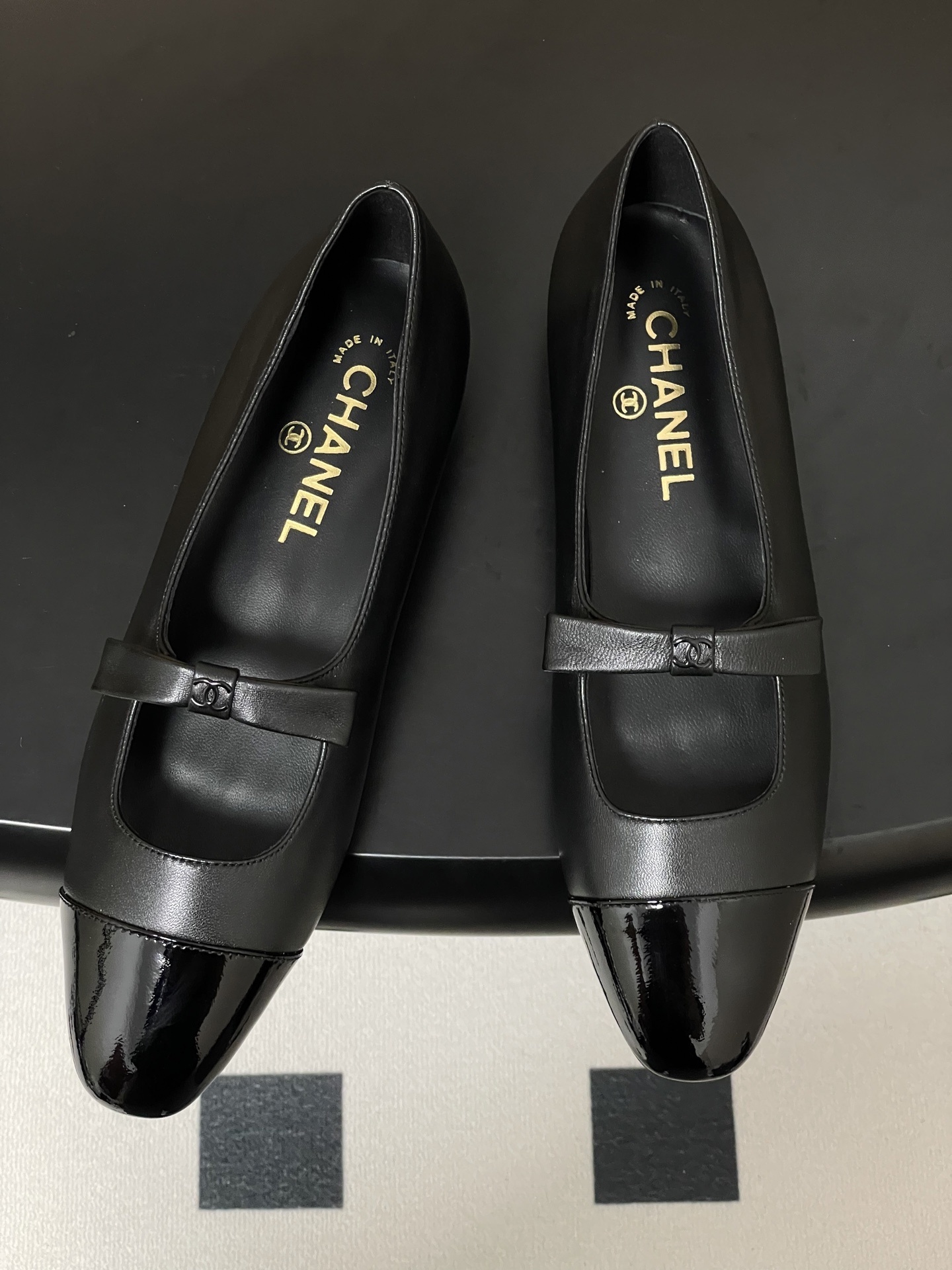 A pair of authentic Chanel black leather Mary Jane flats with glossy patent leather toe caps and CC logo straps displayed on a black surface. - Hình 2