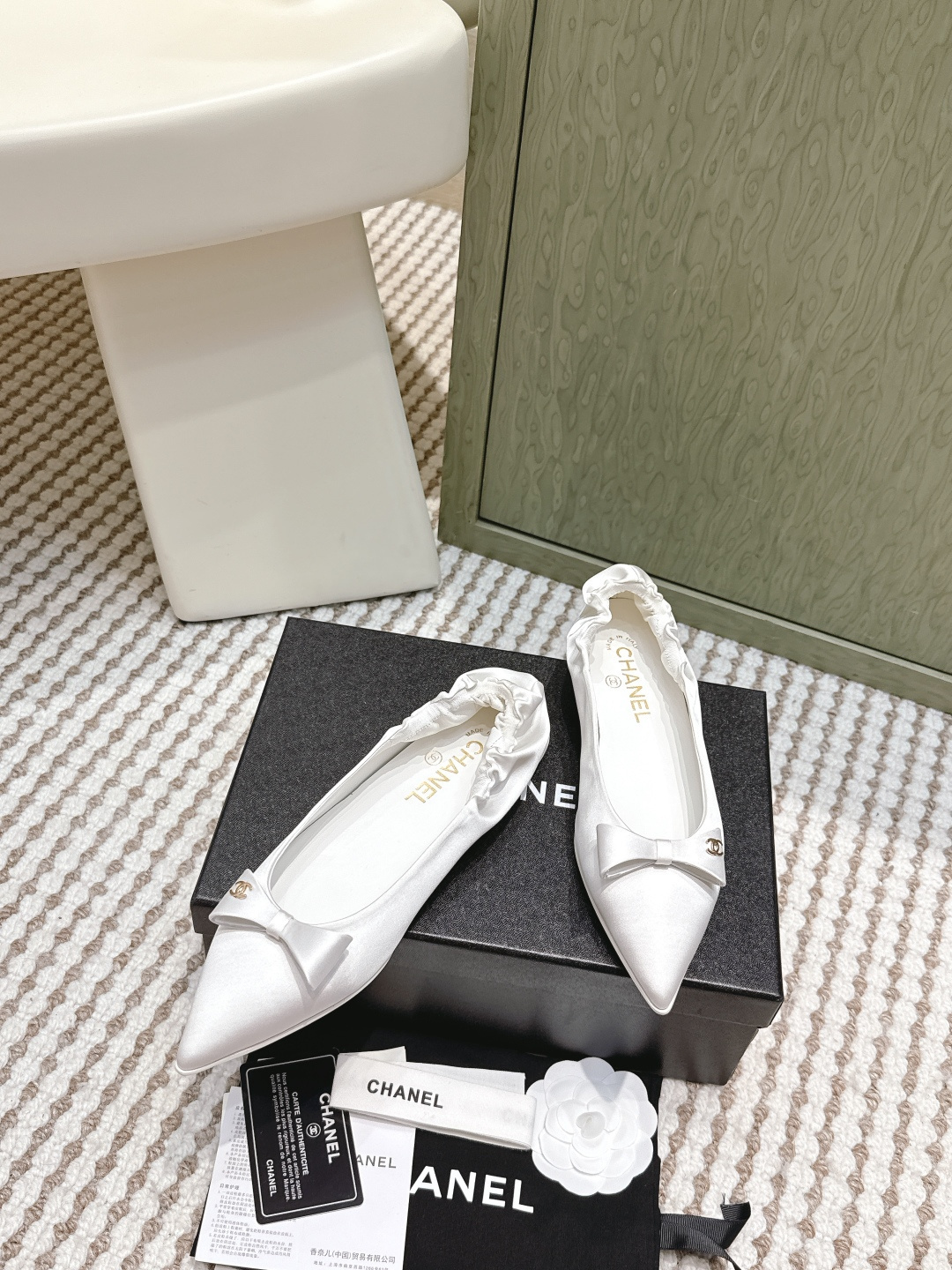 Pair of elegant Chanel white satin pointed-toe ballerina flats with bows and gold logos on a black box. - Hình 3