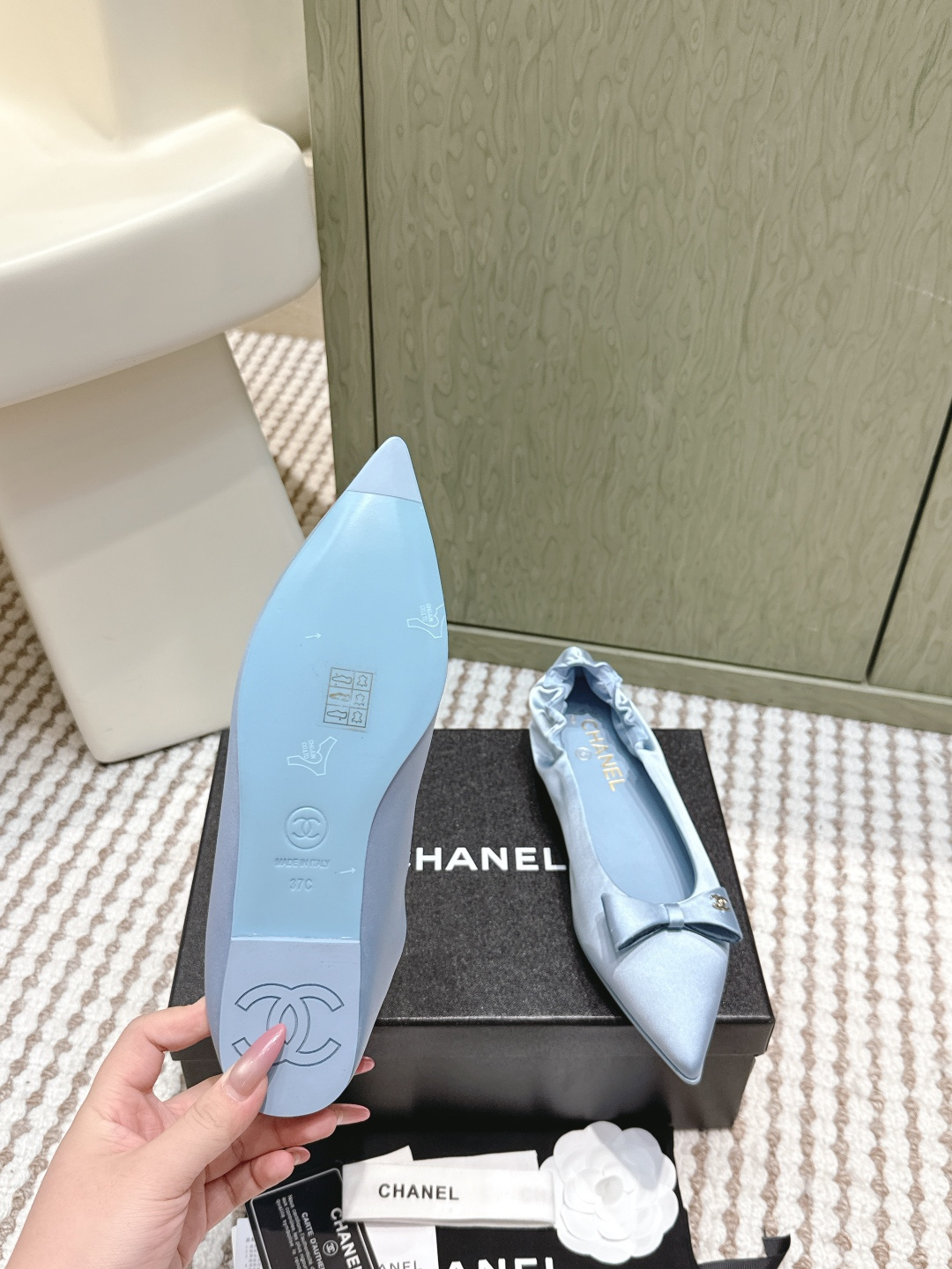 A pair of light blue Chanel satin pointed-toe ballet flats with a bow and gold logo detail, sitting on a black Chanel shoe box. - Hình 9