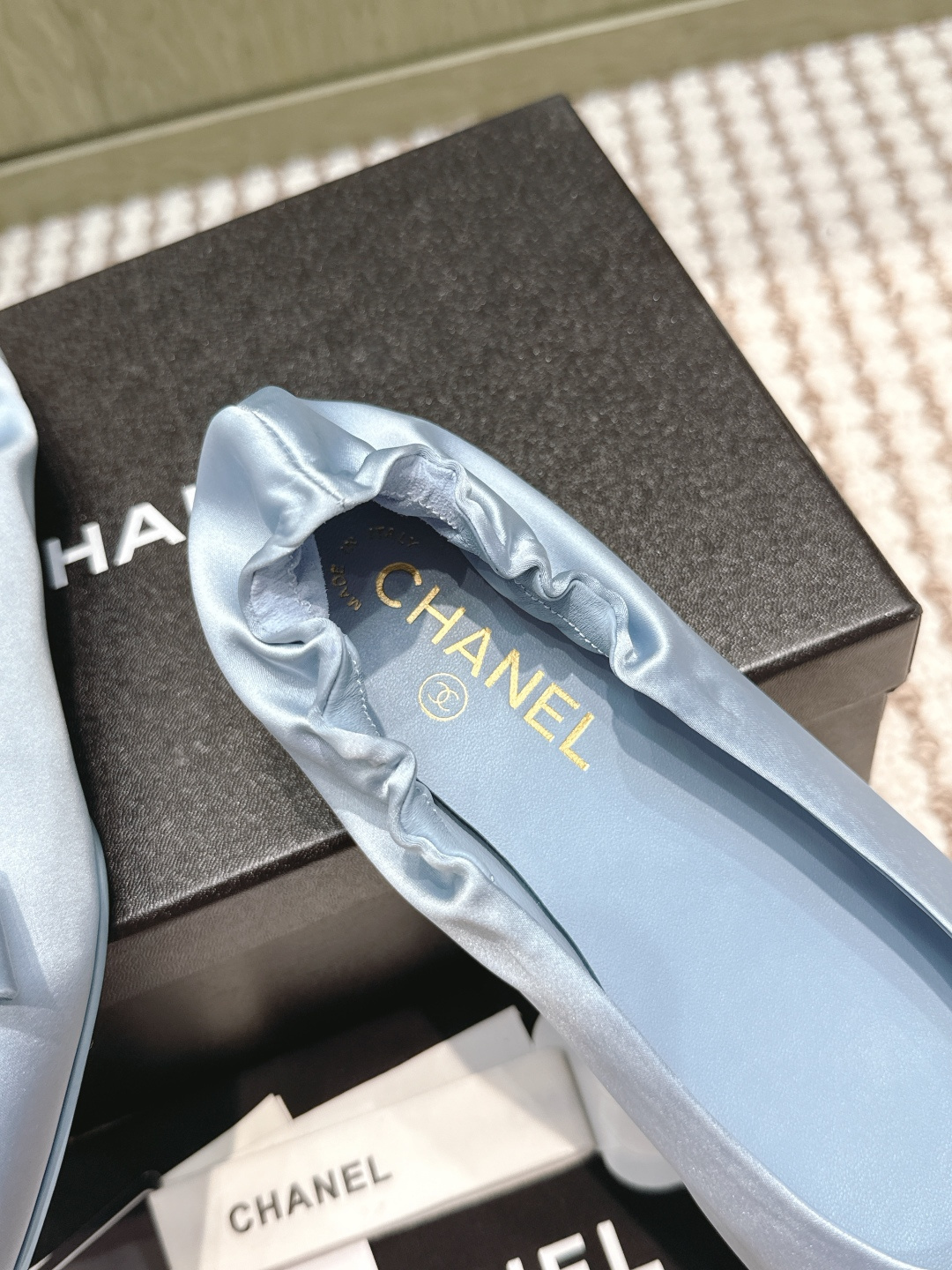 A pair of light blue Chanel satin pointed-toe ballet flats with a bow and gold logo detail, sitting on a black Chanel shoe box. - Hình 7