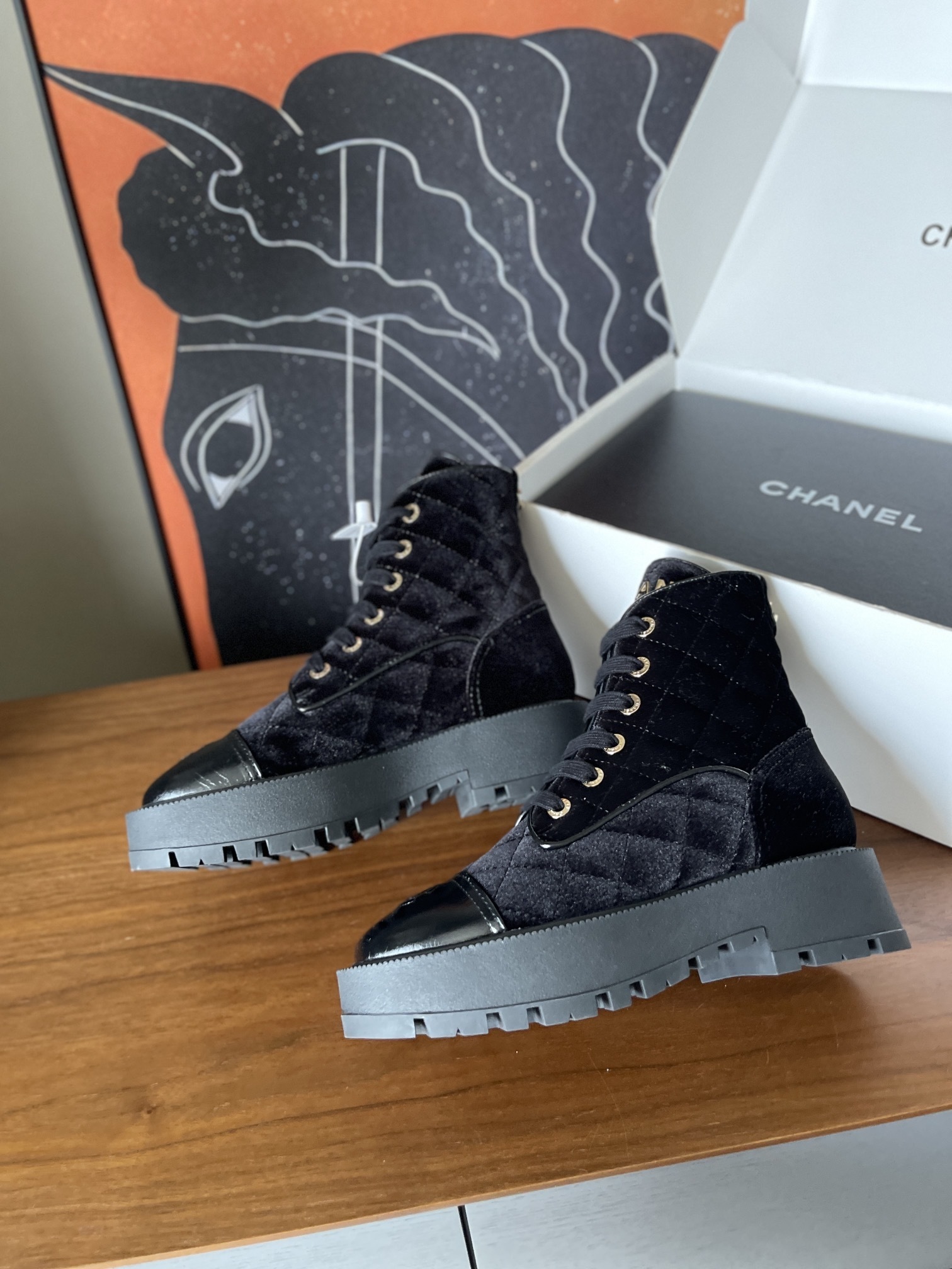 A pair of black Chanel quilted velvet combat boots with patent leather toe caps and gold lettering on a wooden surface. - Hình 4