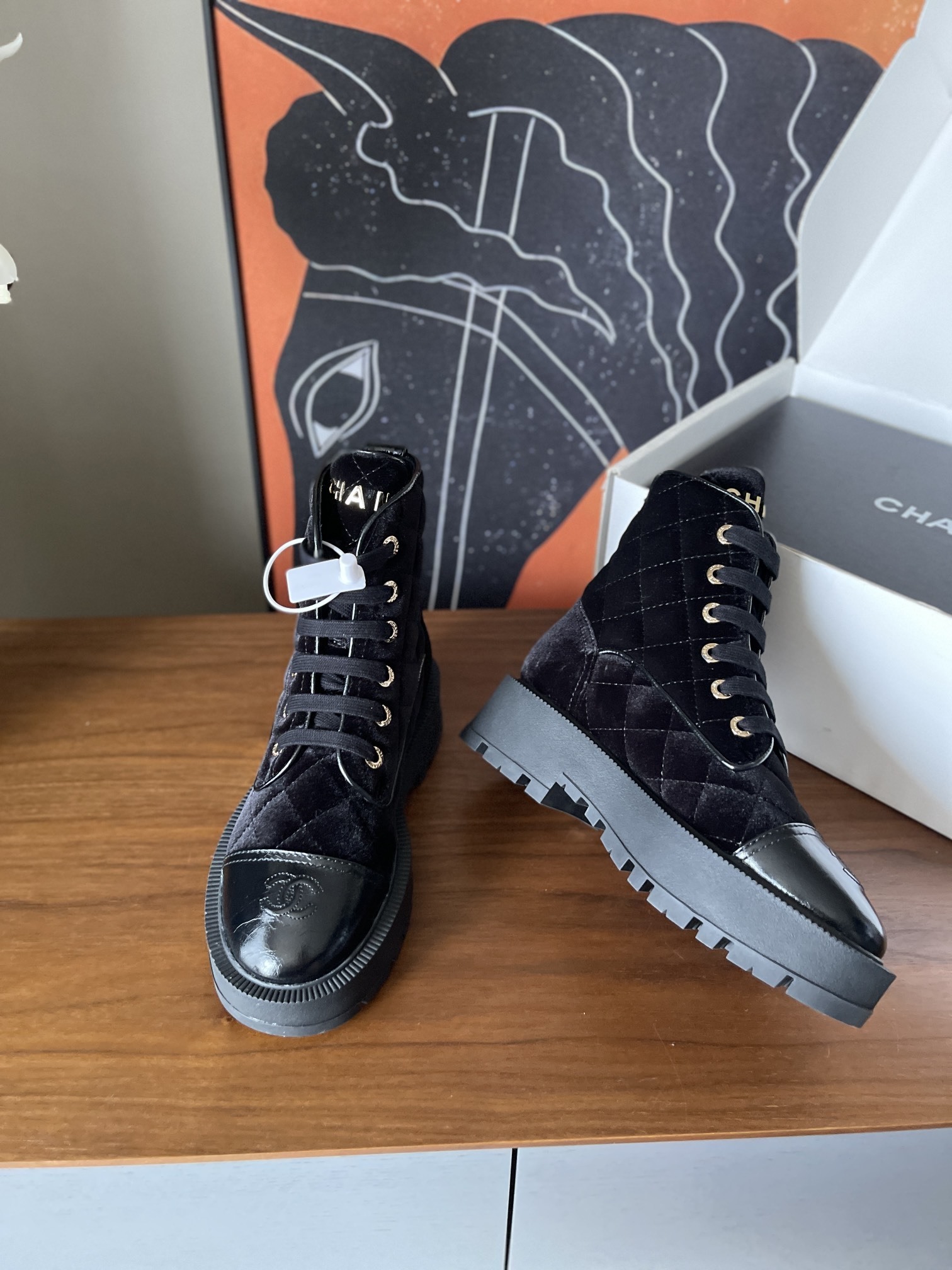 A pair of black Chanel quilted velvet combat boots with patent leather toe caps and gold lettering on a wooden surface. - Hình 3