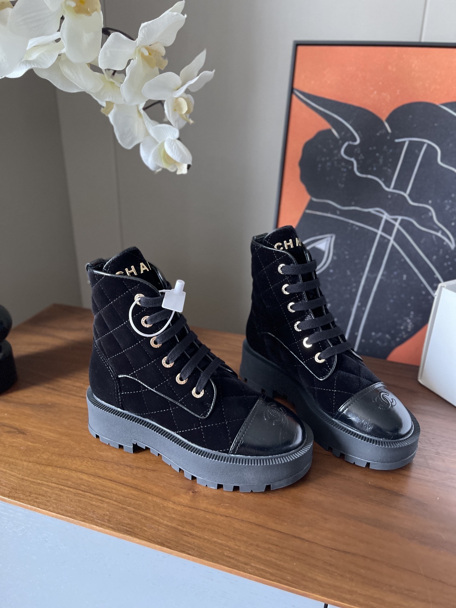 A pair of black Chanel quilted velvet combat boots with patent leather toe caps and gold lettering on a wooden surface. - Hình 2