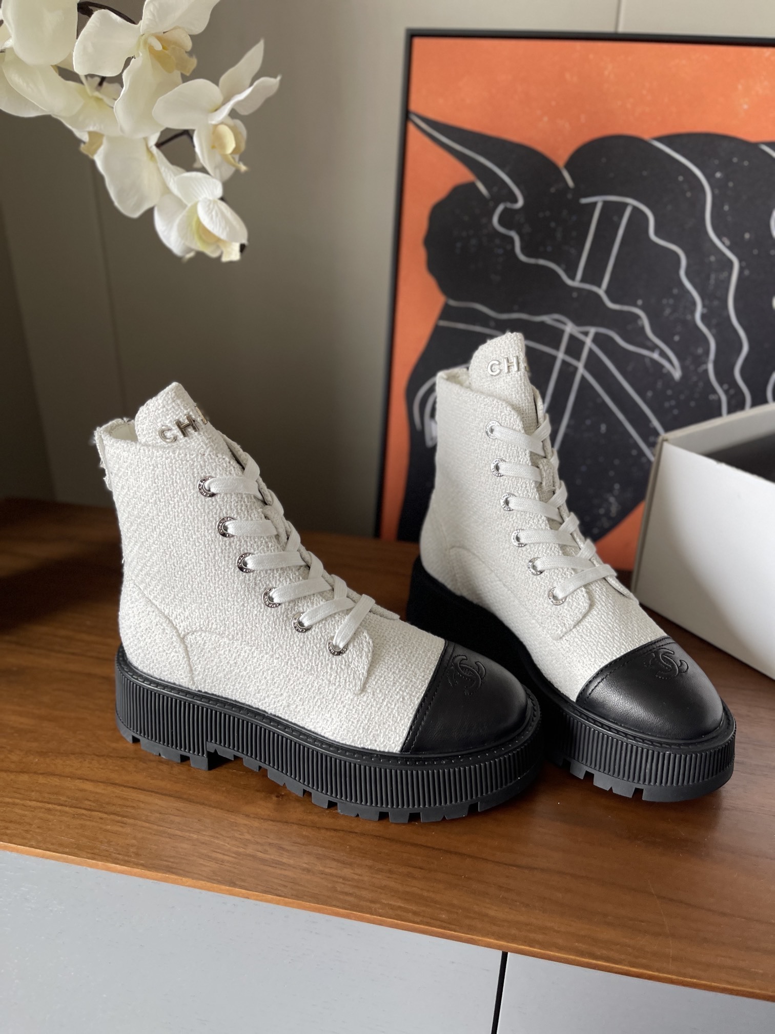 Pair of Chanel white tweed ankle boots with black leather toe caps and chunky black platform soles on a wooden surface. - Hình 2