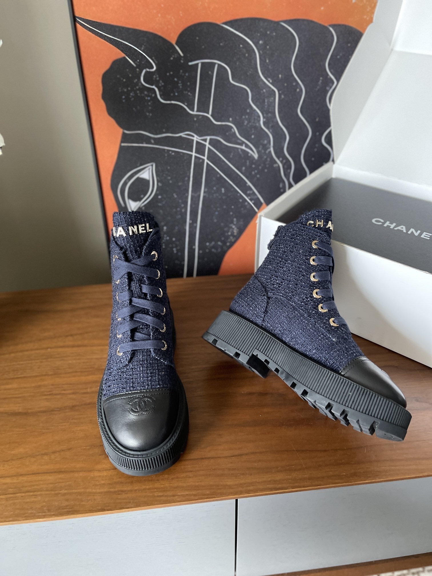 Pair of Chanel navy blue tweed lace-up ankle boots with black leather toe caps and gold lettering on a wooden surface. - Hình 5