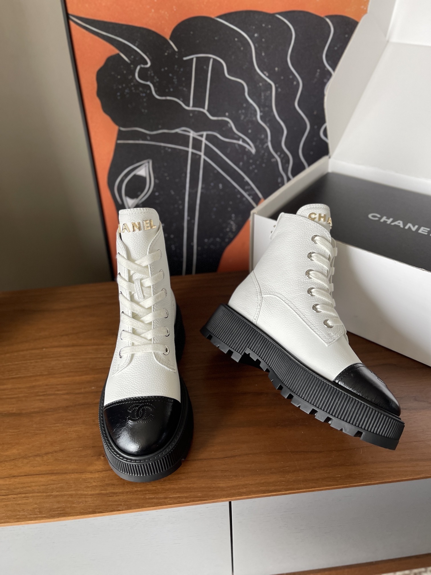 Pair of luxury white leather Chanel ankle boots with black toe caps and chunky platform soles on a wooden table. - Hình 4