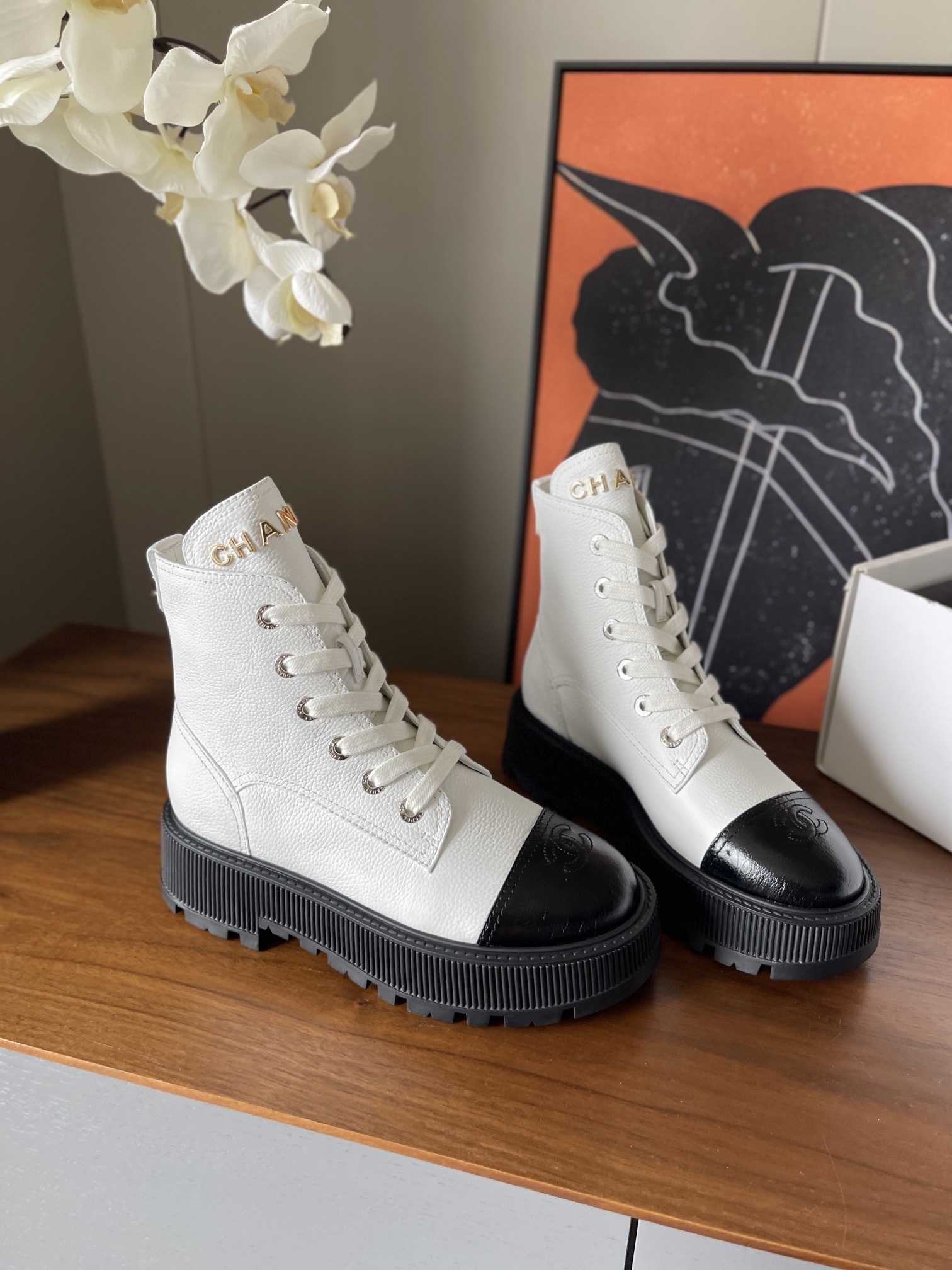 Pair of luxury white leather Chanel ankle boots with black toe caps and chunky platform soles on a wooden table. - Hình 3