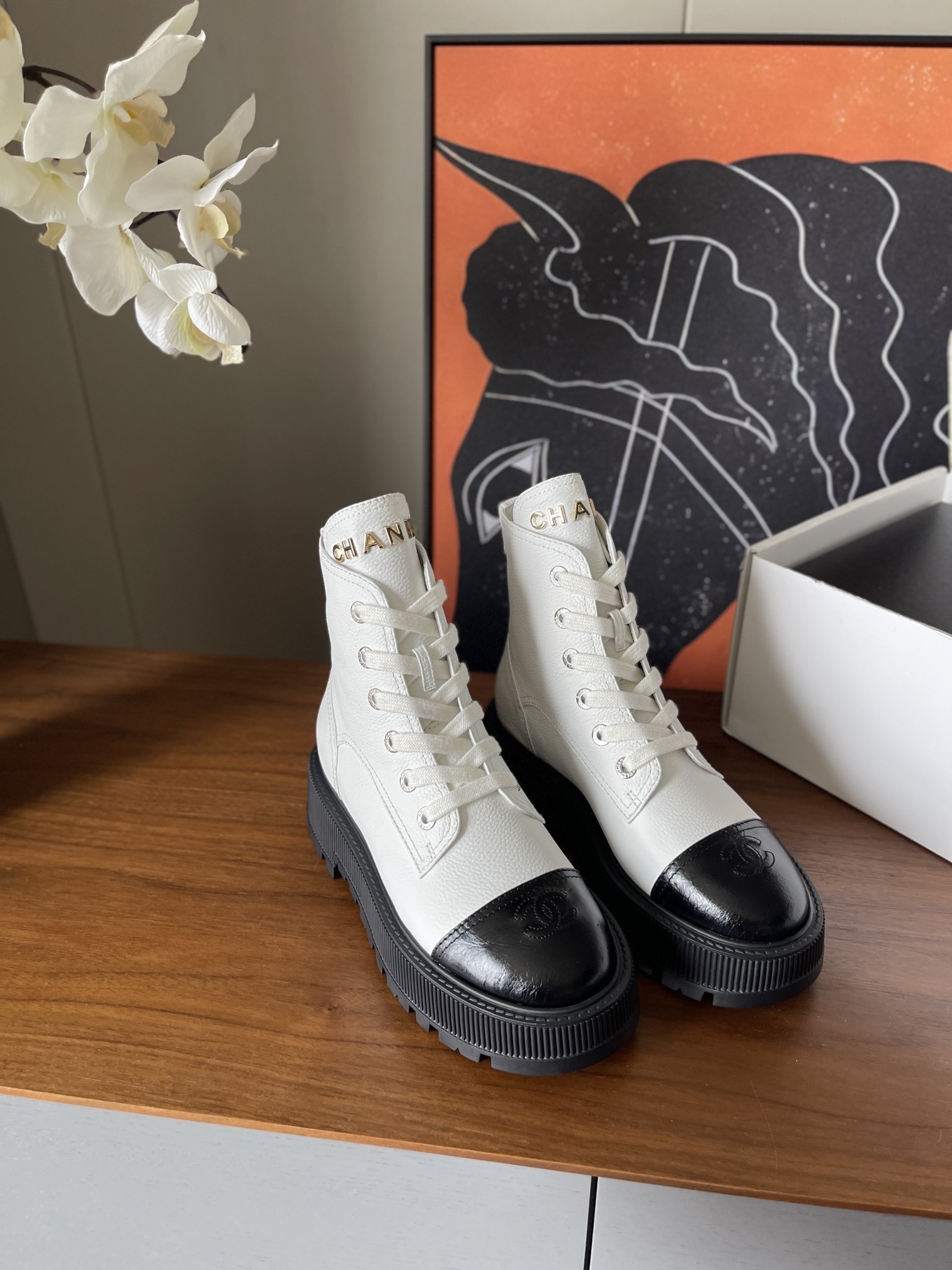 Pair of luxury white leather Chanel ankle boots with black toe caps and chunky platform soles on a wooden table. - Hình 2