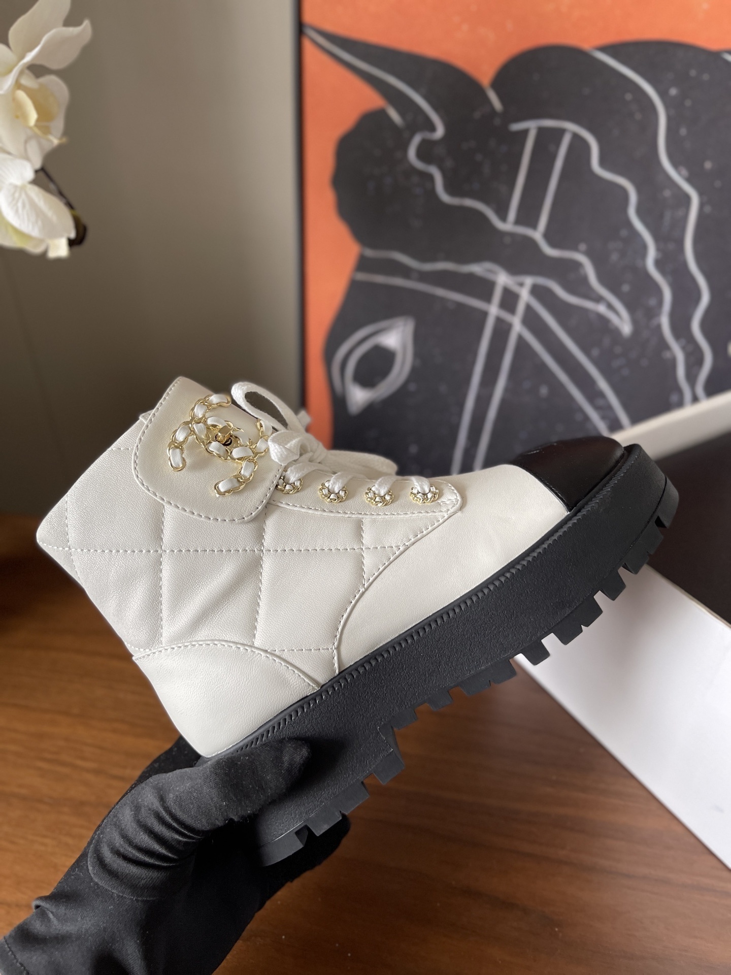 Pair of white quilted ankle boots with black cap toes and gold chain logo detail on a wooden surface - Hình 5