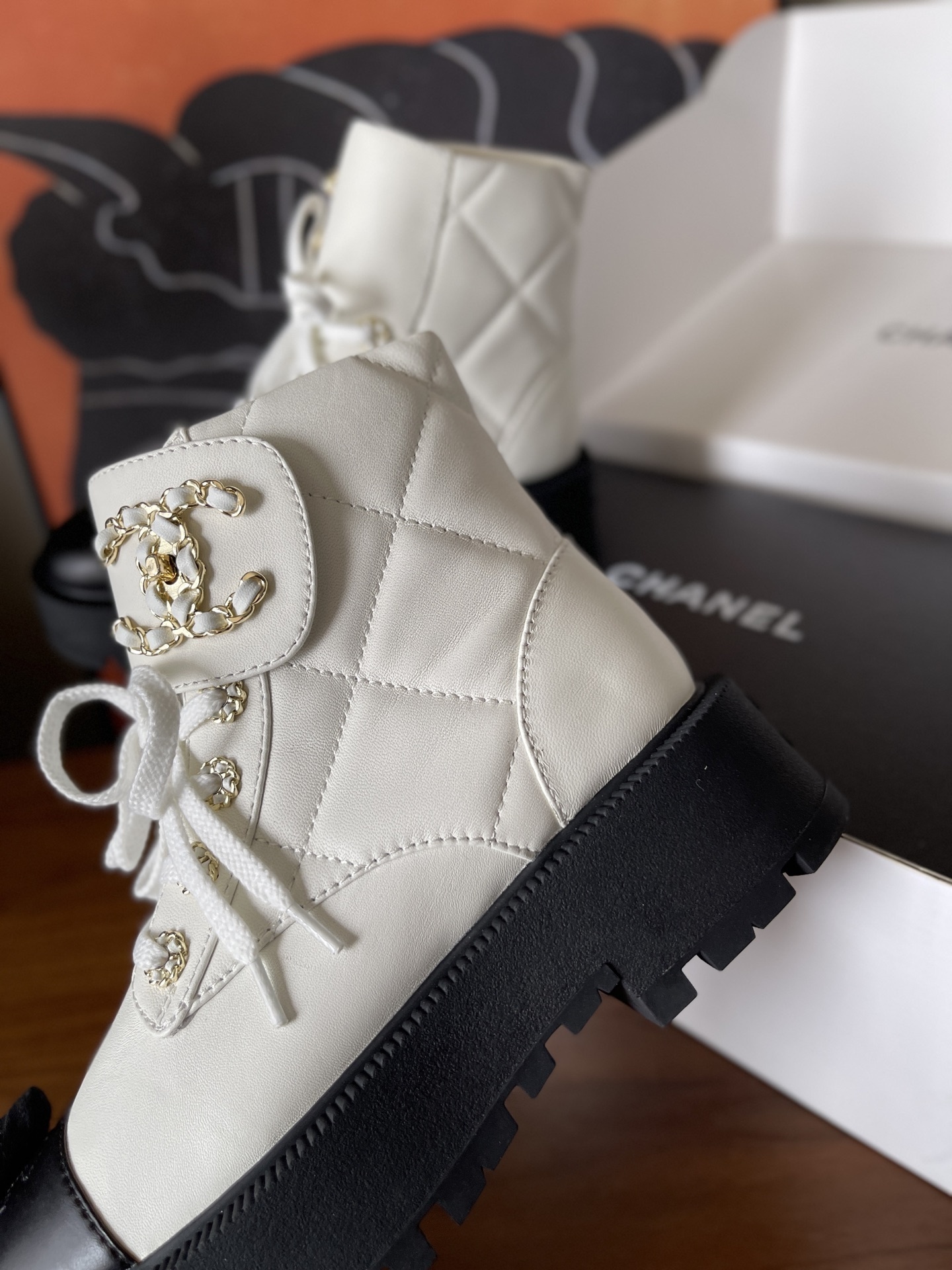 Pair of white quilted ankle boots with black cap toes and gold chain logo detail on a wooden surface - Hình 4