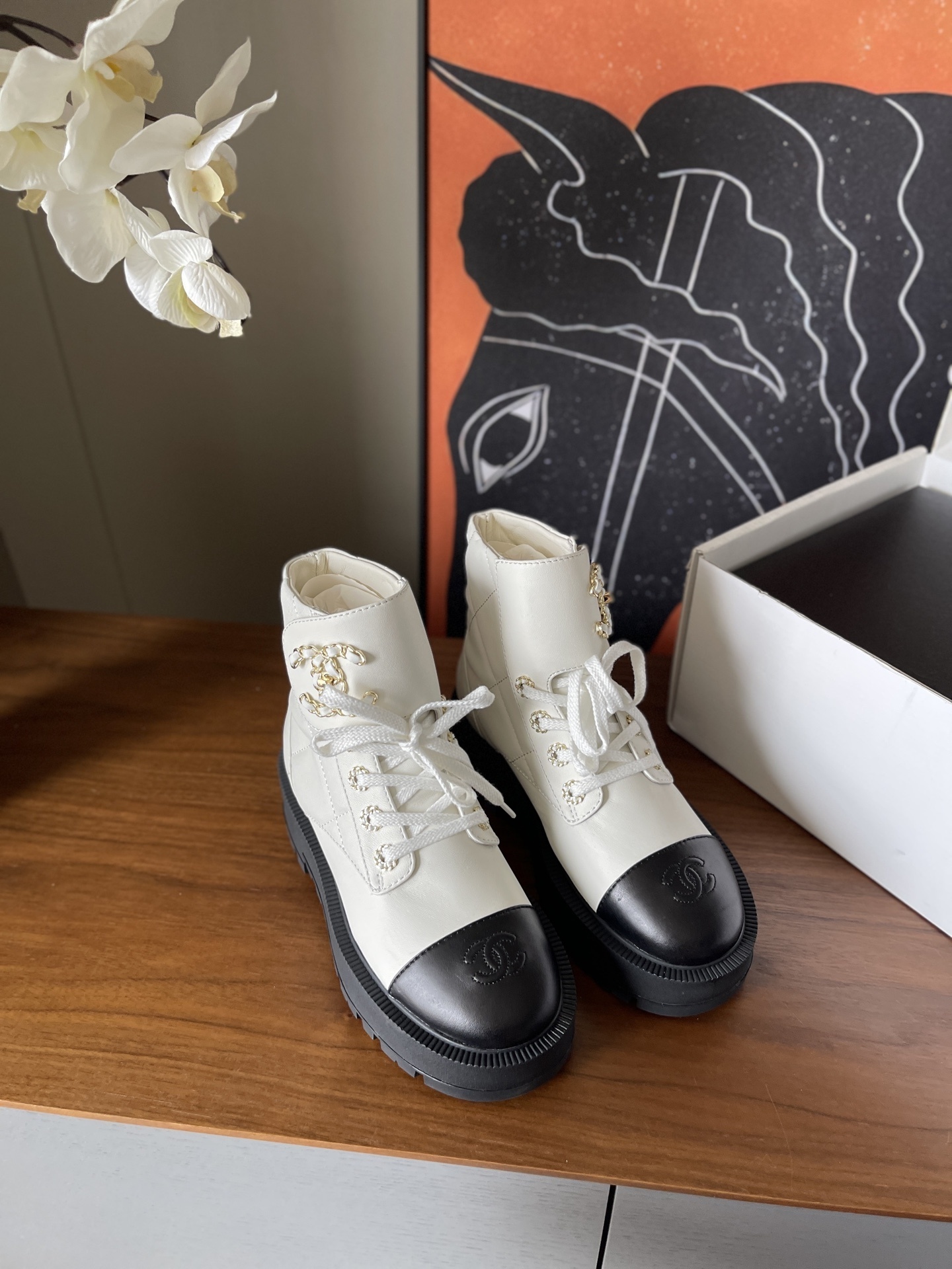 Pair of white quilted ankle boots with black cap toes and gold chain logo detail on a wooden surface - Hình 2