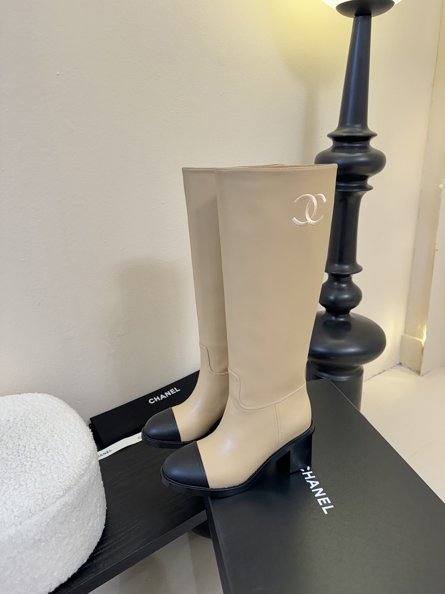 Chanel beige leather knee-high boots with black cap toes and white CC logo on a black display stand - Hình 5