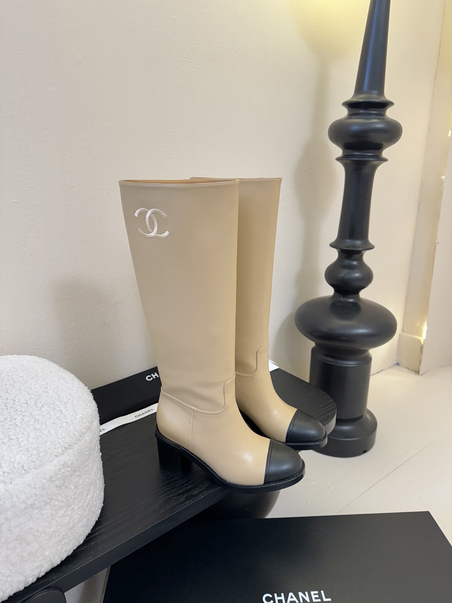 Chanel beige leather knee-high boots with black cap toes and white CC logo on a black display stand - Hình 2