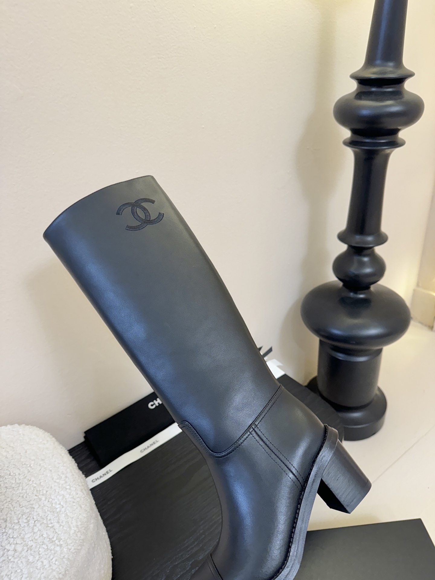 A pair of elegant Chanel black calfskin leather knee-high boots with an embroidered CC logo and block heels displayed on a black stand. - Hình 8