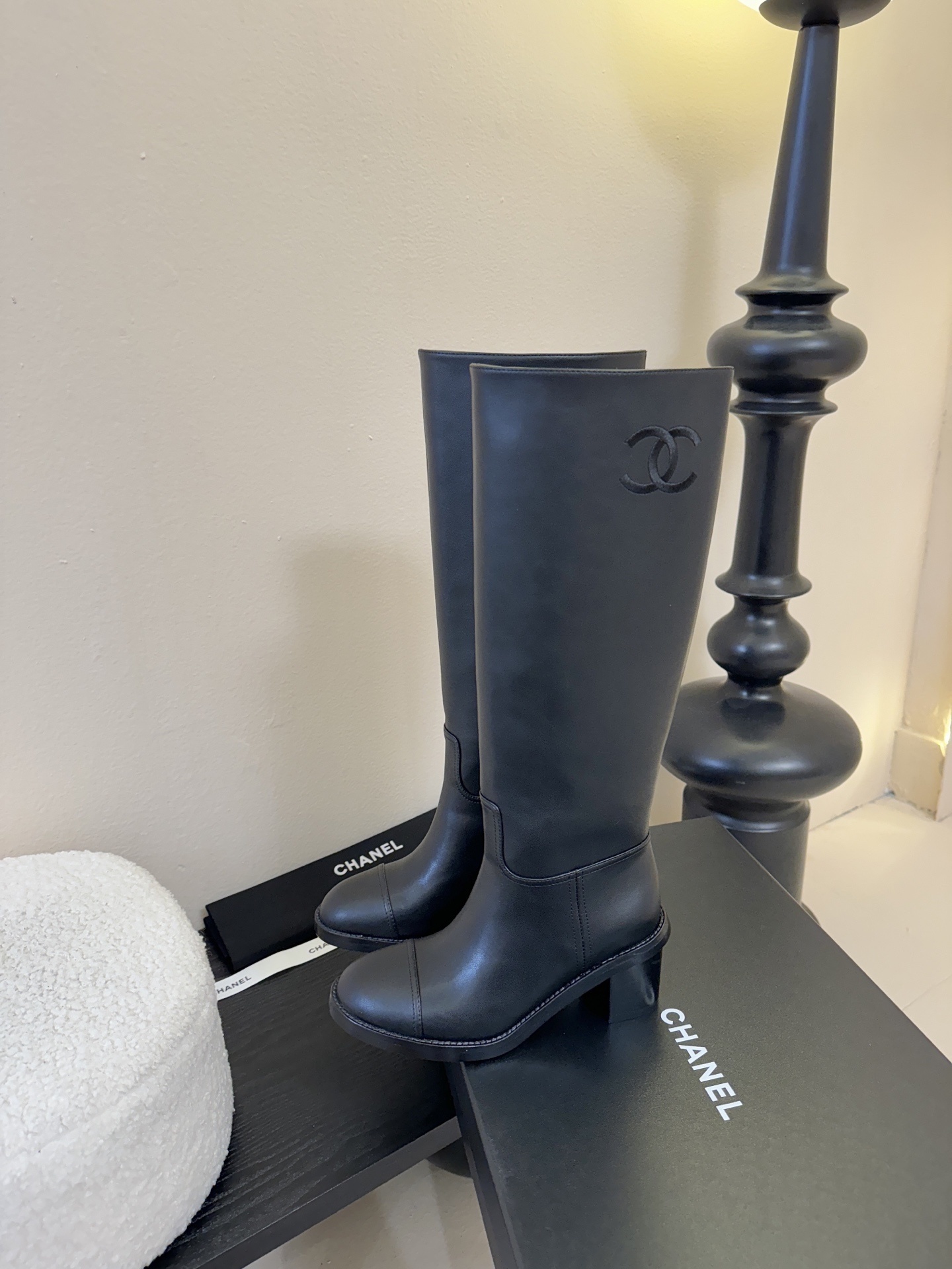 A pair of elegant Chanel black calfskin leather knee-high boots with an embroidered CC logo and block heels displayed on a black stand. - Hình 5