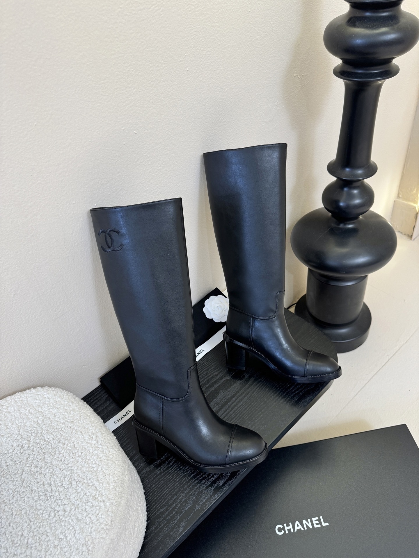 A pair of elegant Chanel black calfskin leather knee-high boots with an embroidered CC logo and block heels displayed on a black stand. - Hình 4