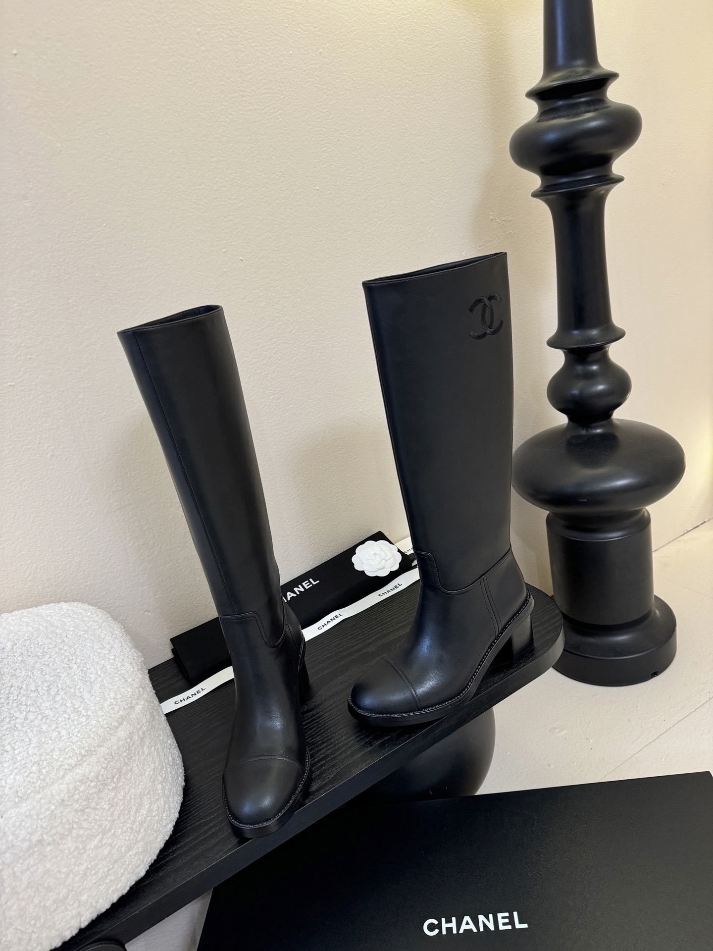 A pair of elegant Chanel black calfskin leather knee-high boots with an embroidered CC logo and block heels displayed on a black stand. - Hình 3