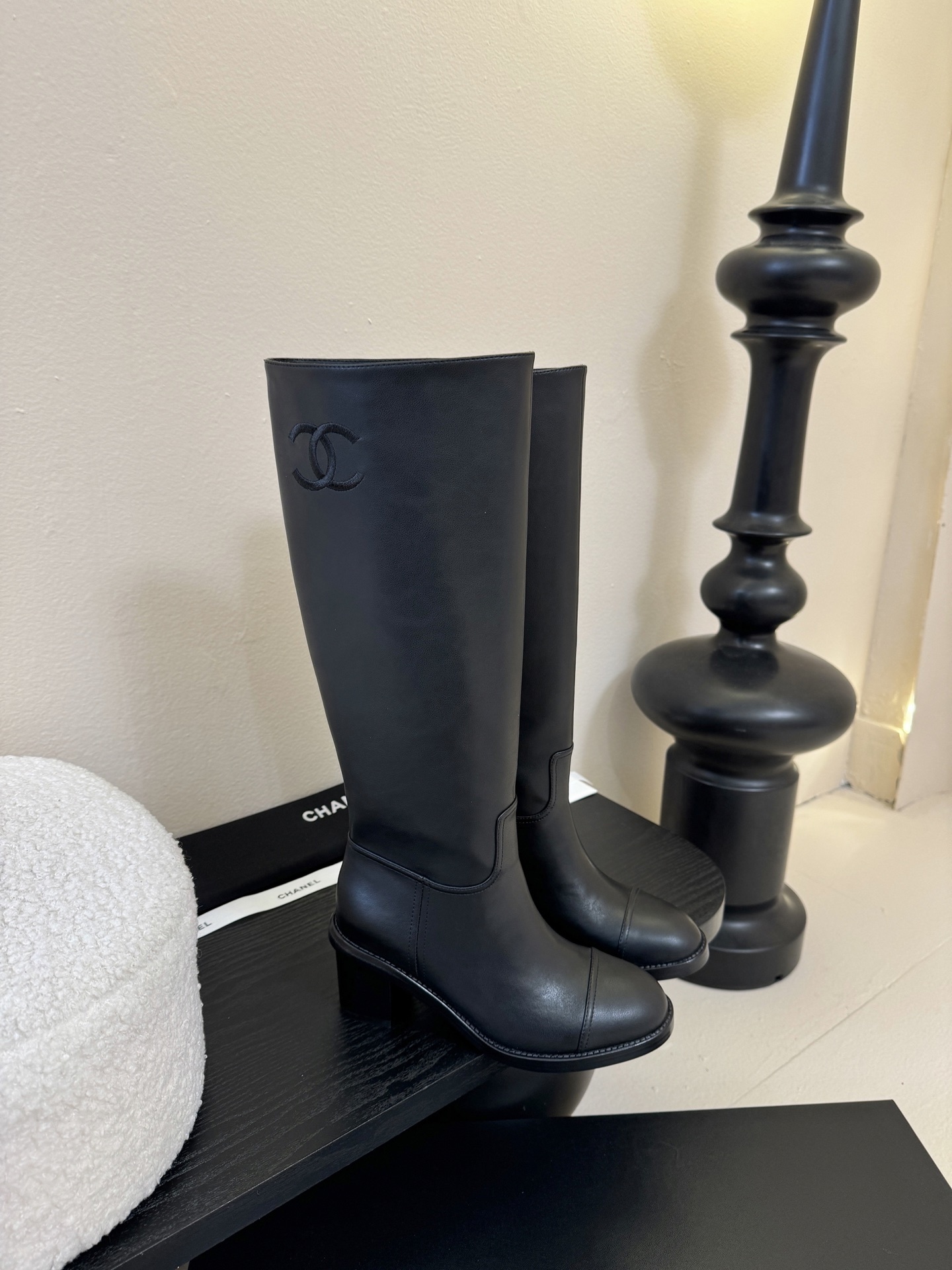 A pair of elegant Chanel black calfskin leather knee-high boots with an embroidered CC logo and block heels displayed on a black stand. - Hình 2
