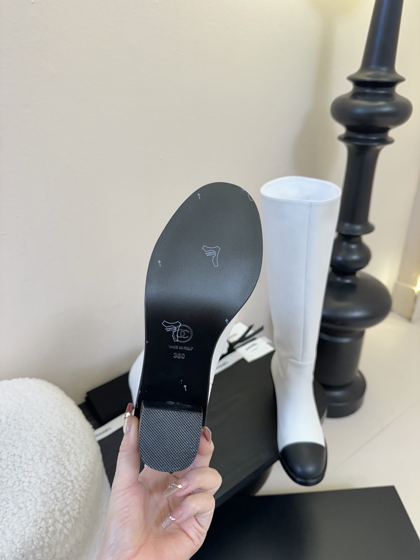 Pair of luxury Chanel white leather knee-high boots with black cap toes and CC logo on a black display stand. - Hình 9