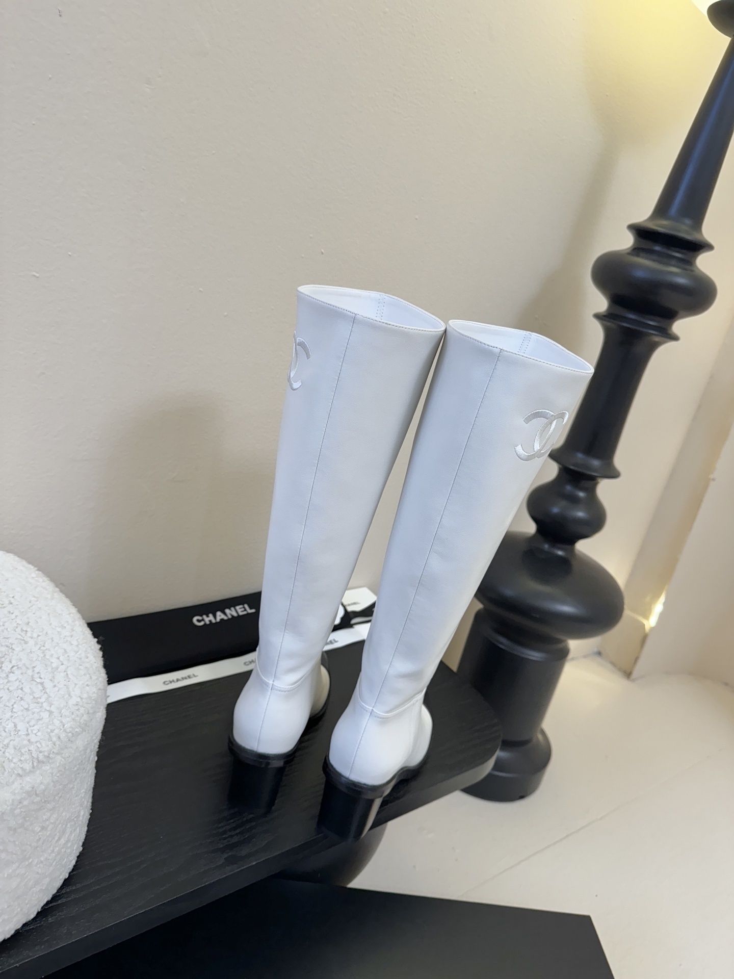 Pair of luxury Chanel white leather knee-high boots with black cap toes and CC logo on a black display stand. - Hình 7