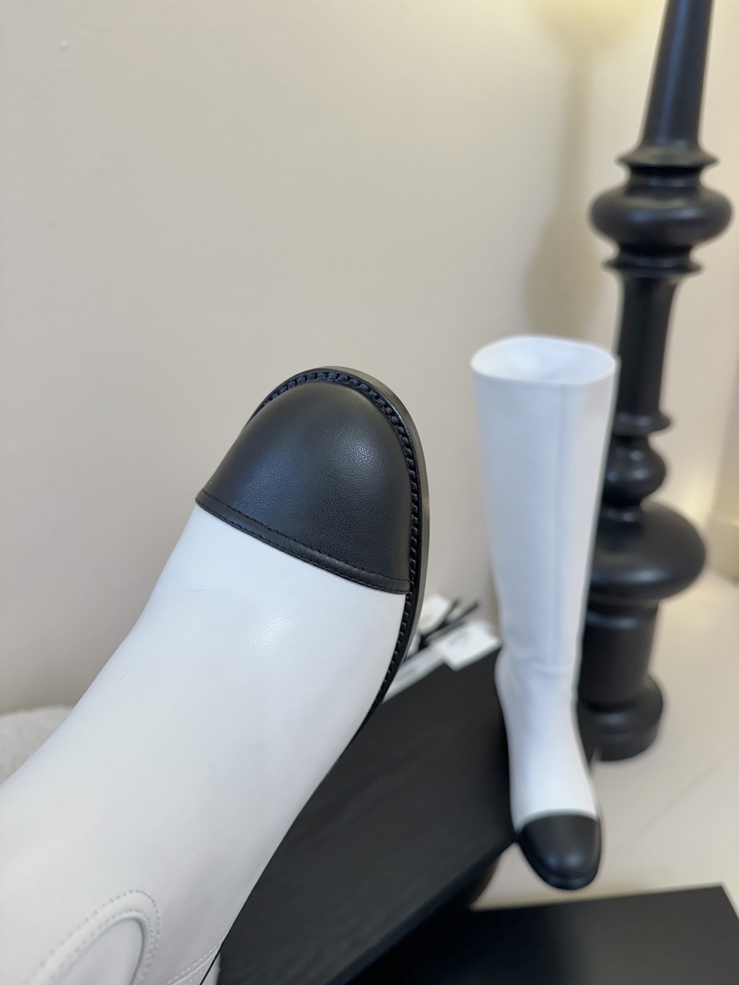 Pair of luxury Chanel white leather knee-high boots with black cap toes and CC logo on a black display stand. - Hình 6