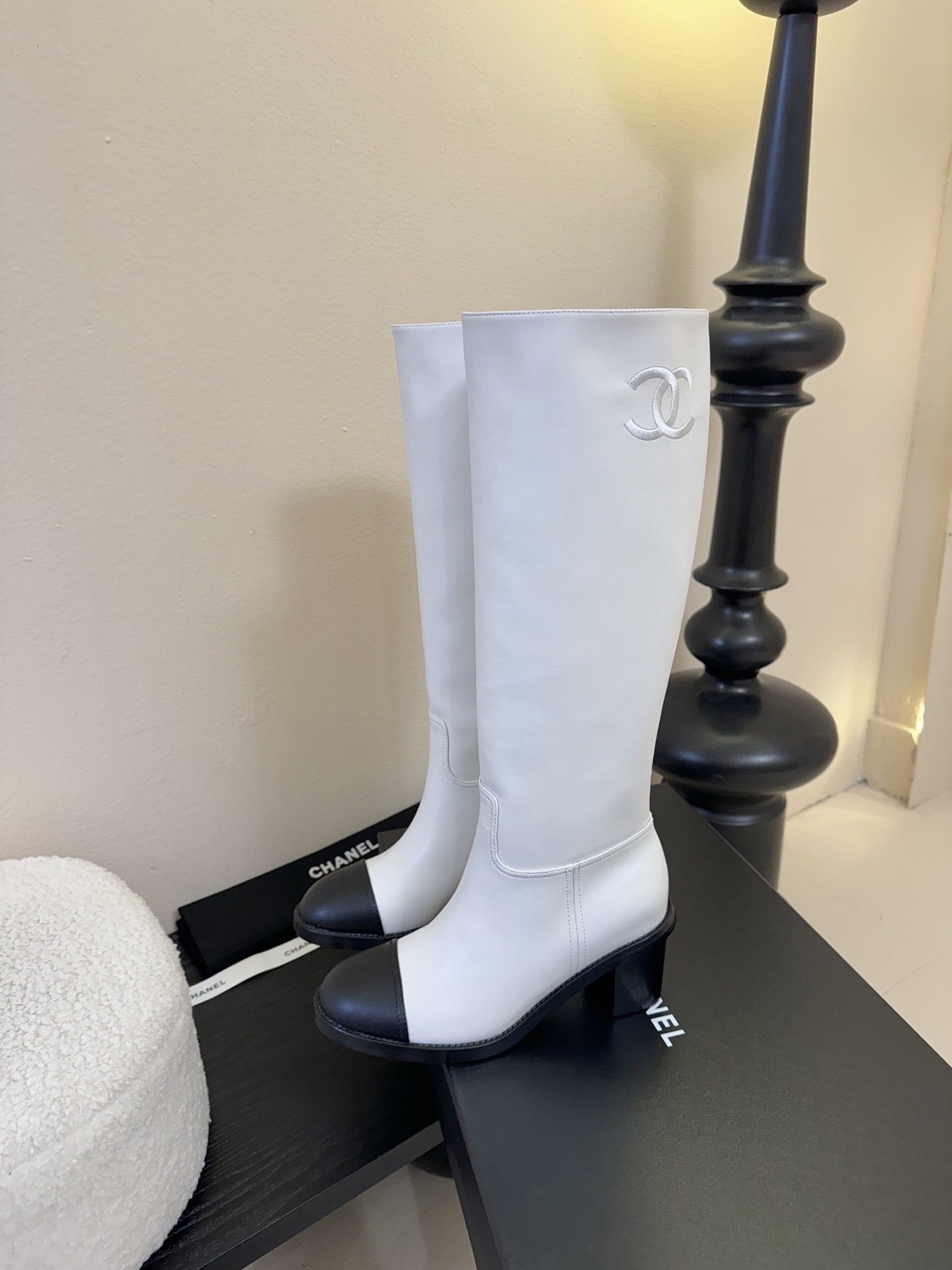 Pair of luxury Chanel white leather knee-high boots with black cap toes and CC logo on a black display stand. - Hình 5