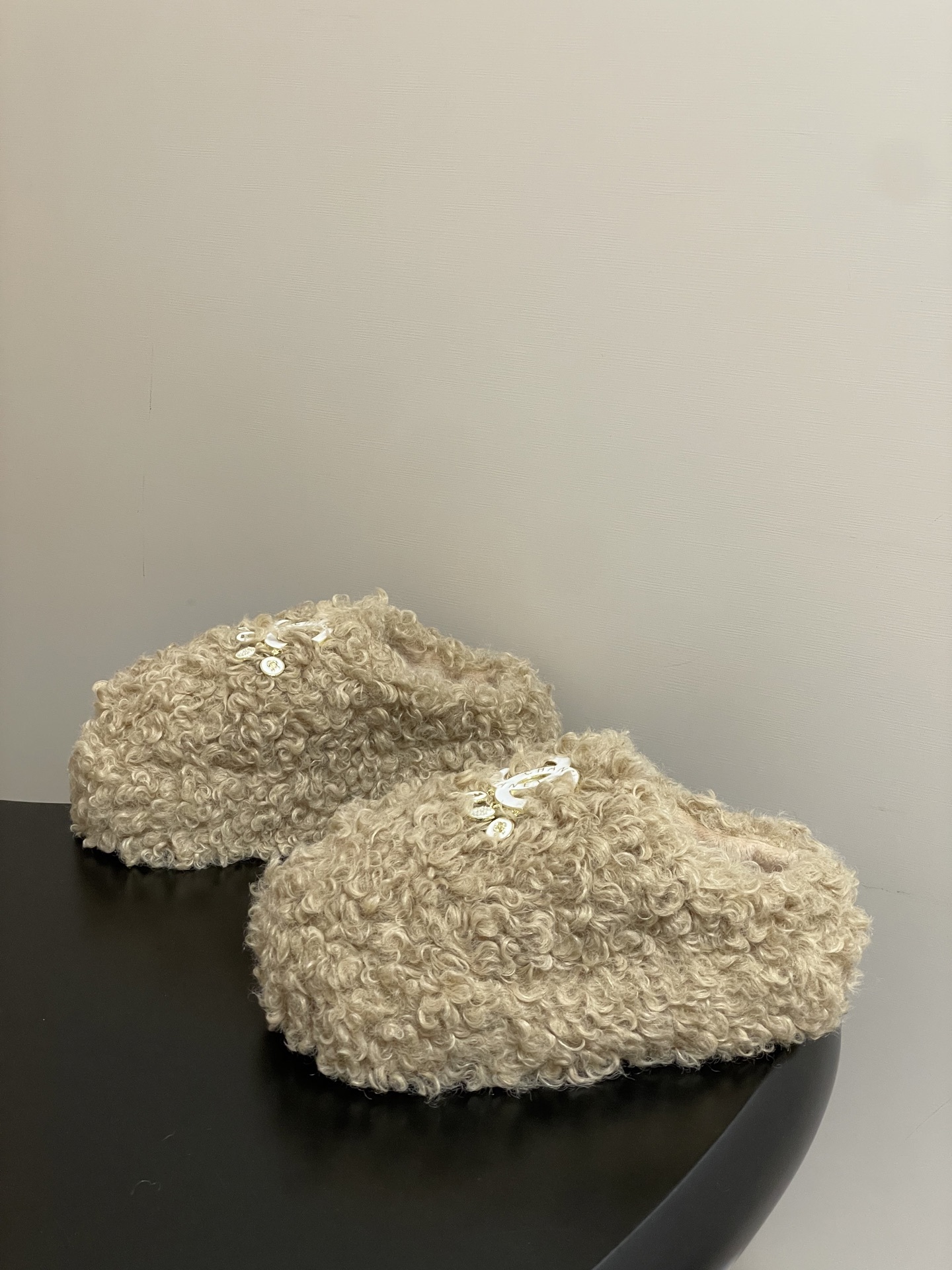 Pair of beige curly faux fur platform slippers featuring a white designer logo with hanging pearl charms on a black surface. - Hình 3