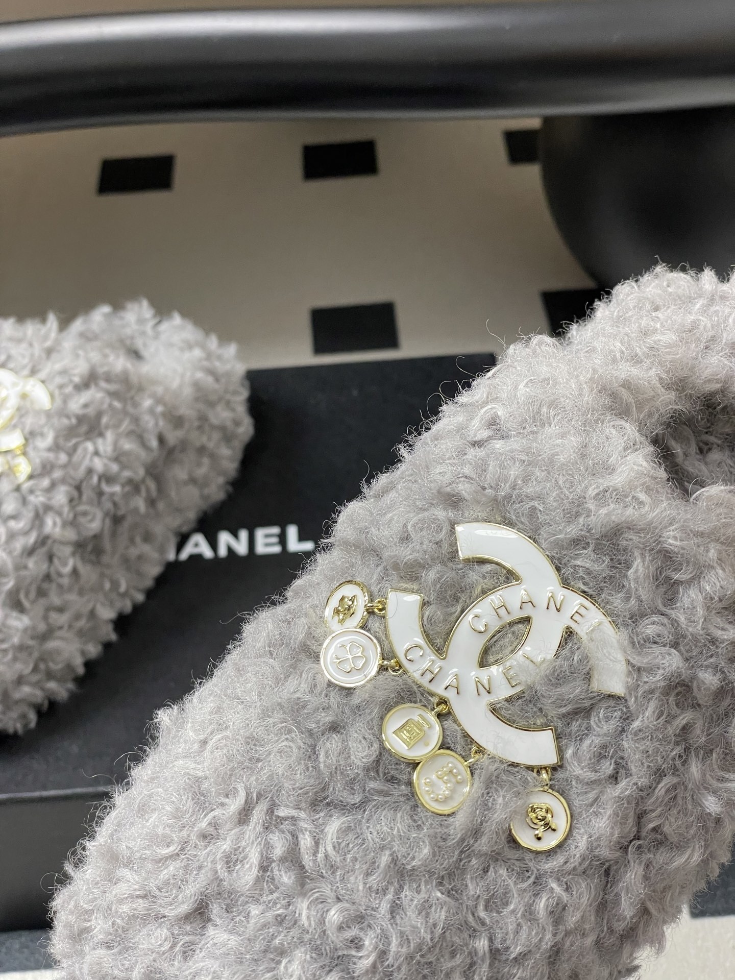 A pair of plush grey curly faux fur slippers featuring a white interlocking logo with gold charms on a black surface. - Hình 9