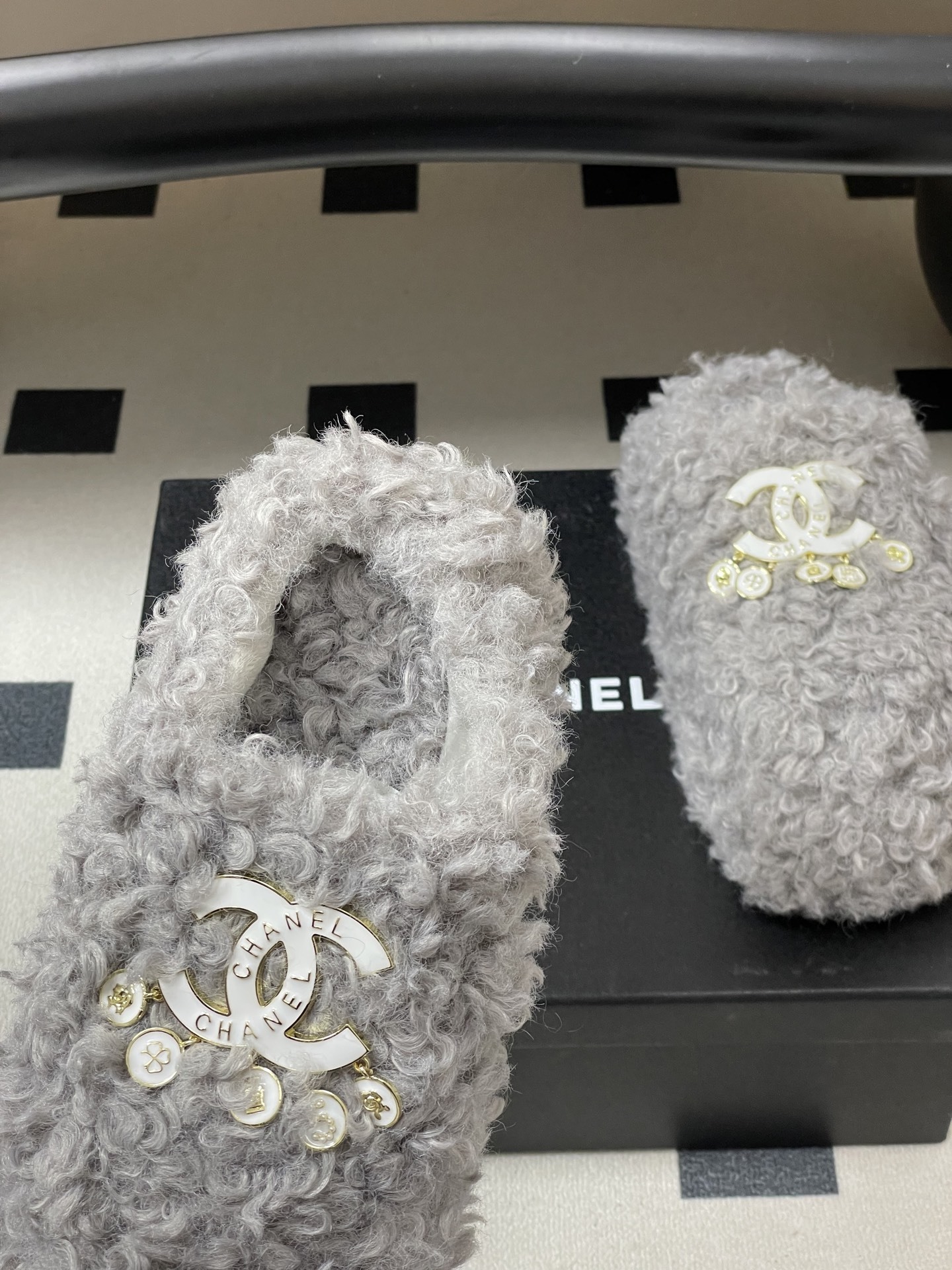 A pair of plush grey curly faux fur slippers featuring a white interlocking logo with gold charms on a black surface. - Hình 7