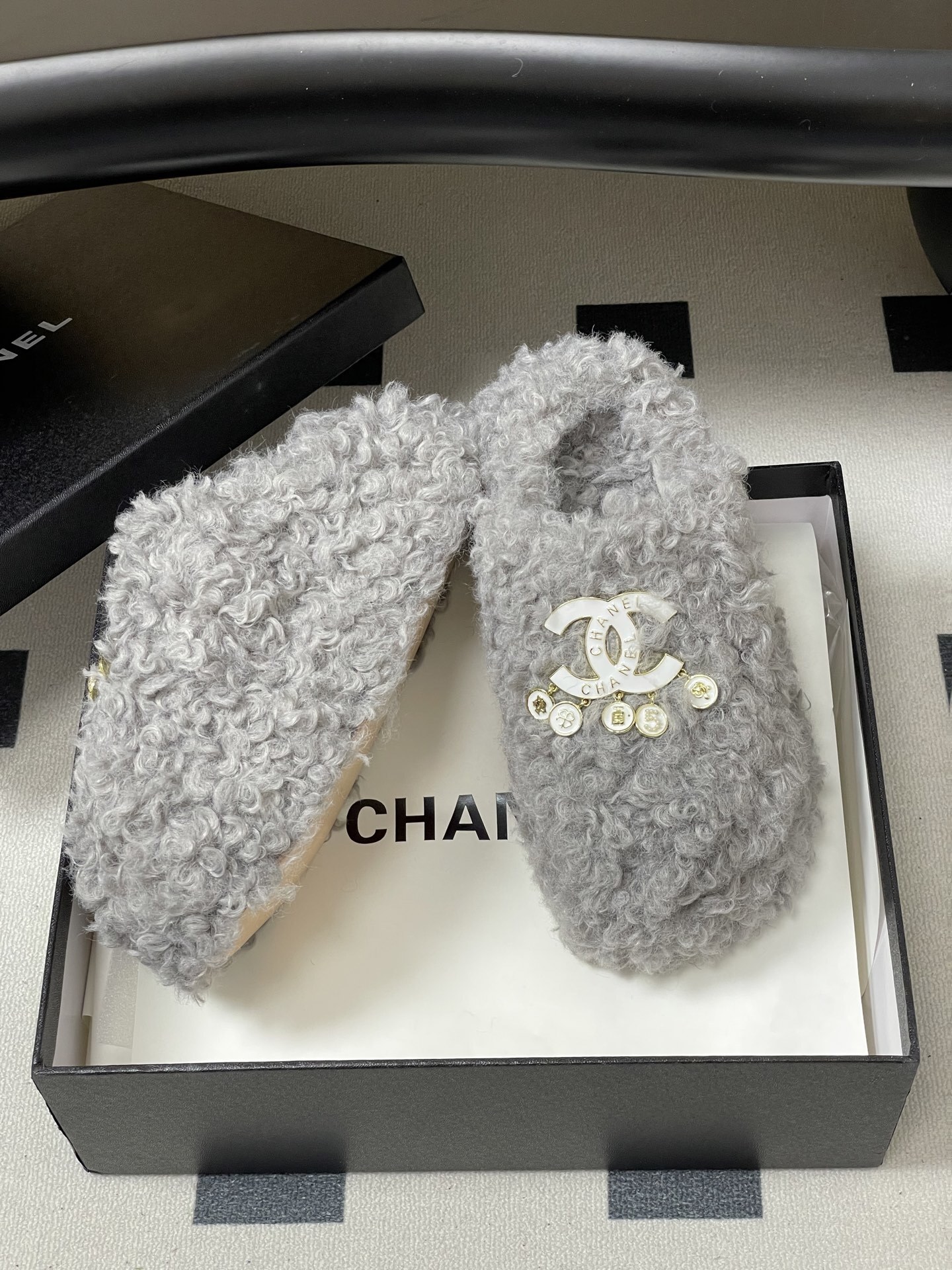 A pair of plush grey curly faux fur slippers featuring a white interlocking logo with gold charms on a black surface. - Hình 6