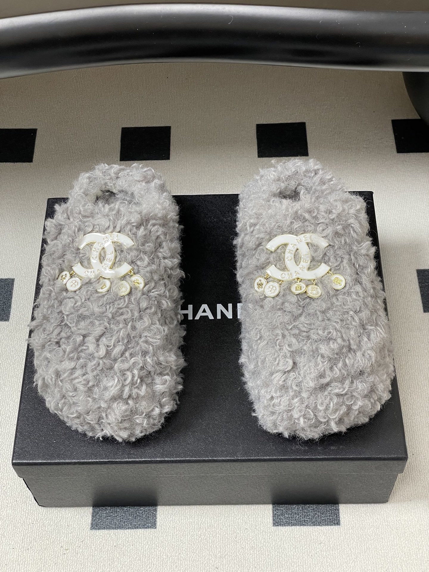 A pair of plush grey curly faux fur slippers featuring a white interlocking logo with gold charms on a black surface. - Hình 5