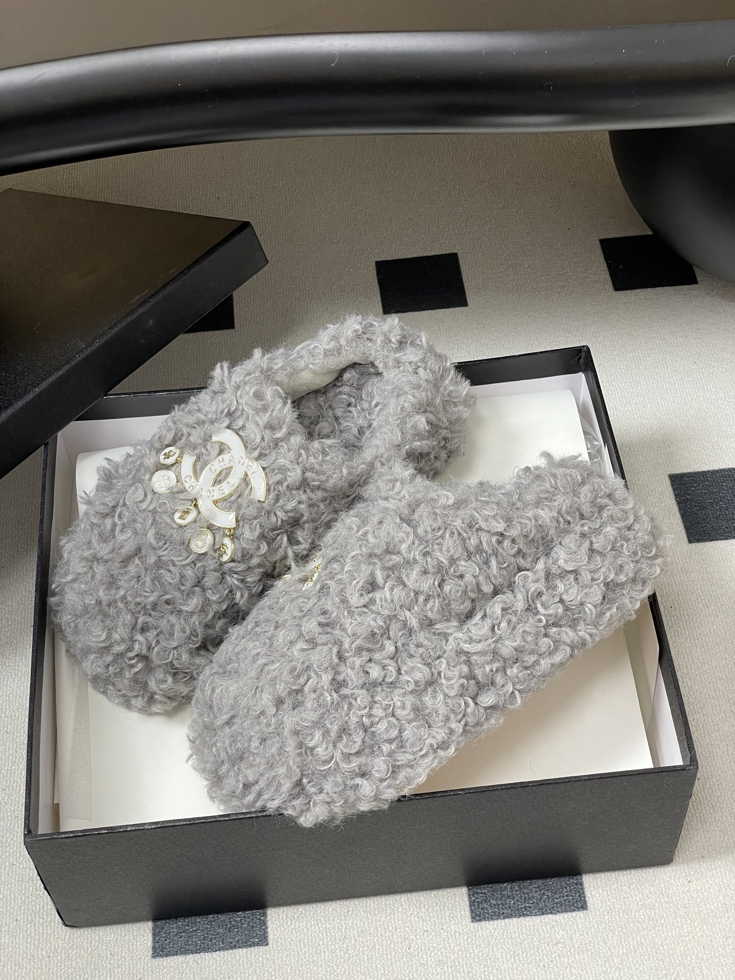 A pair of plush grey curly faux fur slippers featuring a white interlocking logo with gold charms on a black surface. - Hình 4