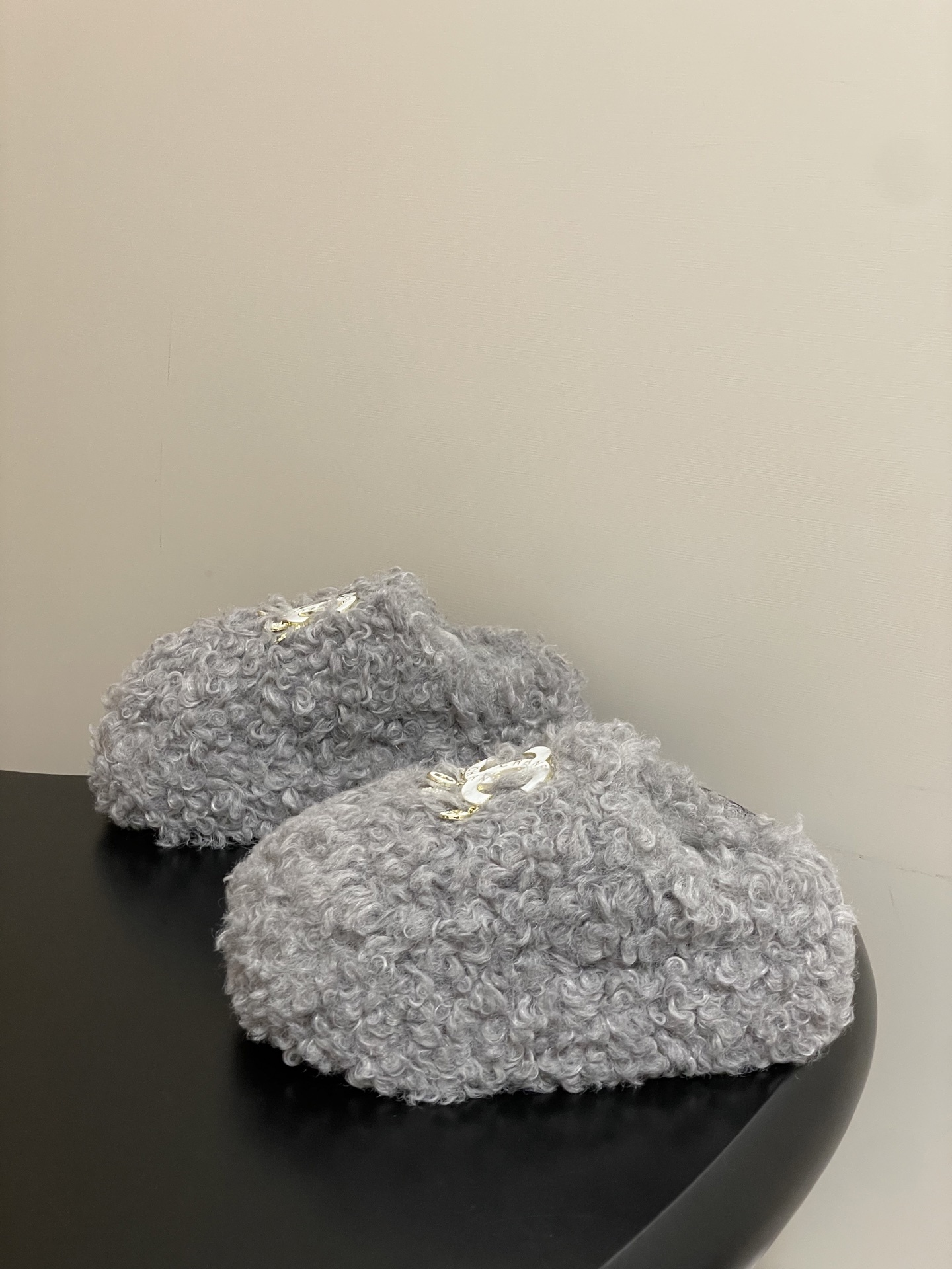 A pair of plush grey curly faux fur slippers featuring a white interlocking logo with gold charms on a black surface. - Hình 3