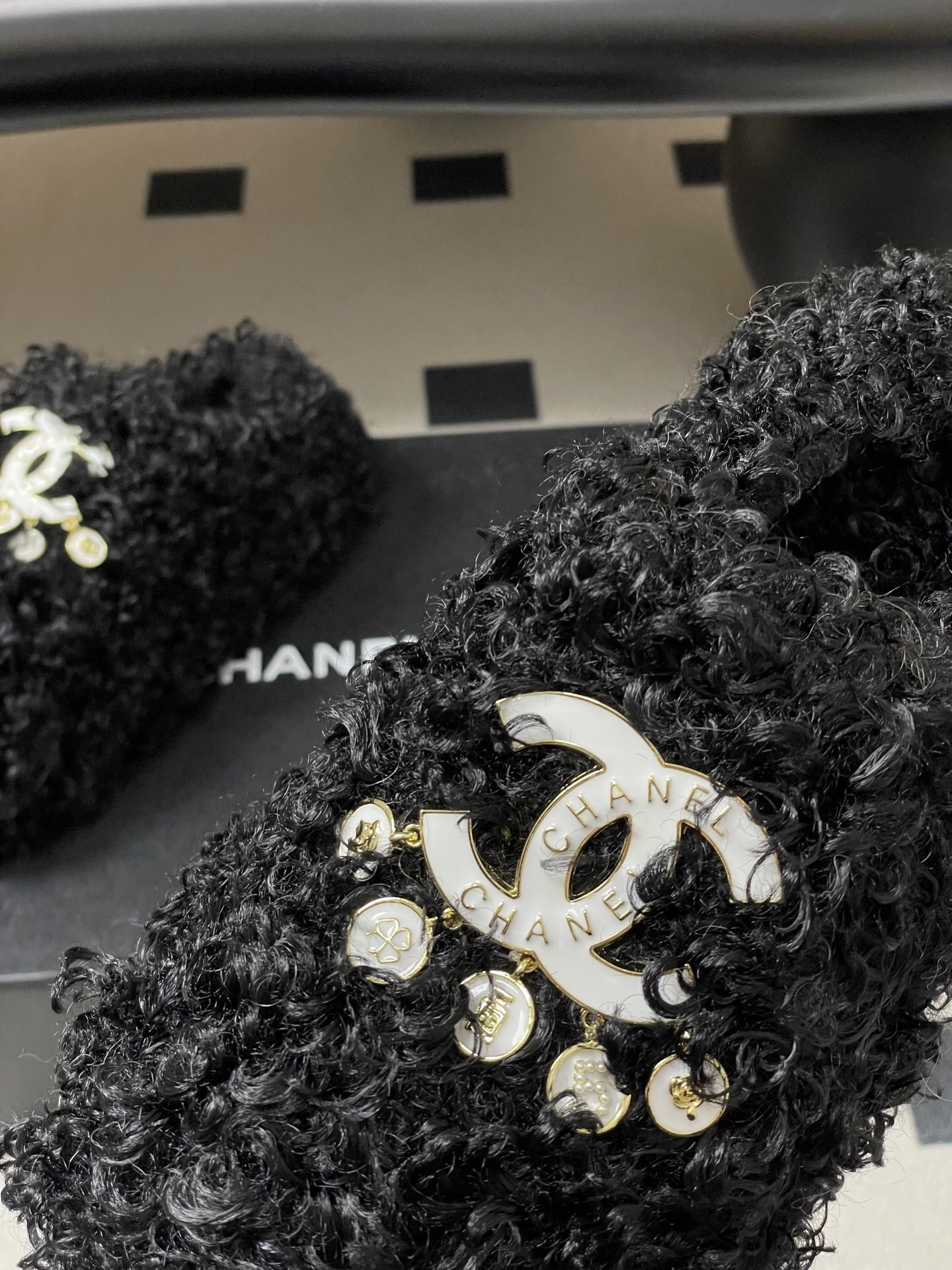 Pair of black curly faux fur loafers with white interlocking logo and dangling charms on a black surface. - Hình 9