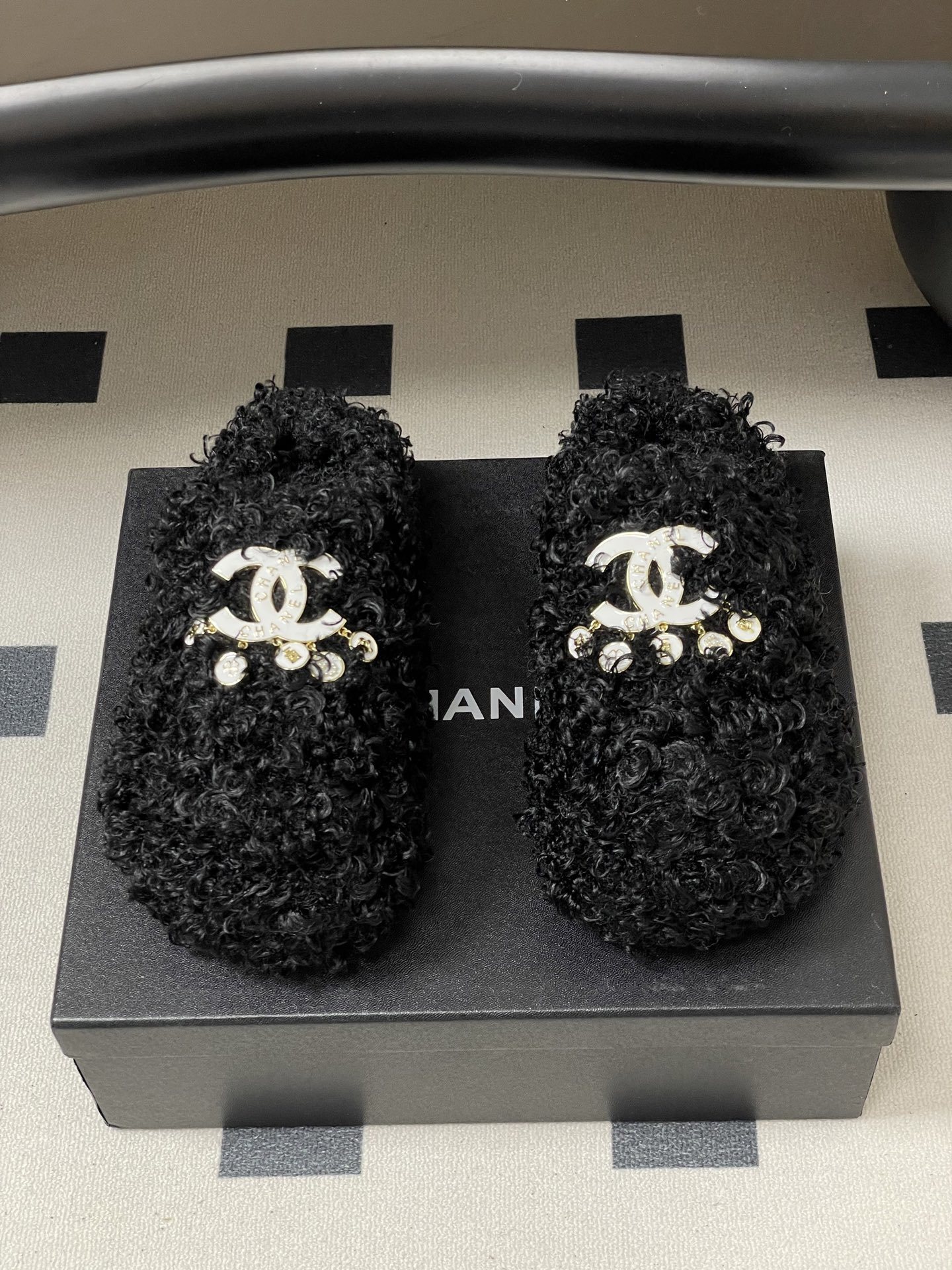 Pair of black curly faux fur loafers with white interlocking logo and dangling charms on a black surface. - Hình 5