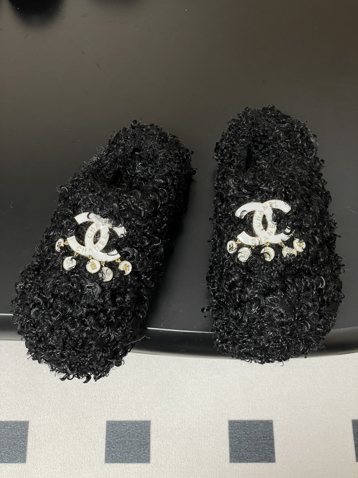 Pair of black curly faux fur loafers with white interlocking logo and dangling charms on a black surface. - Hình 2
