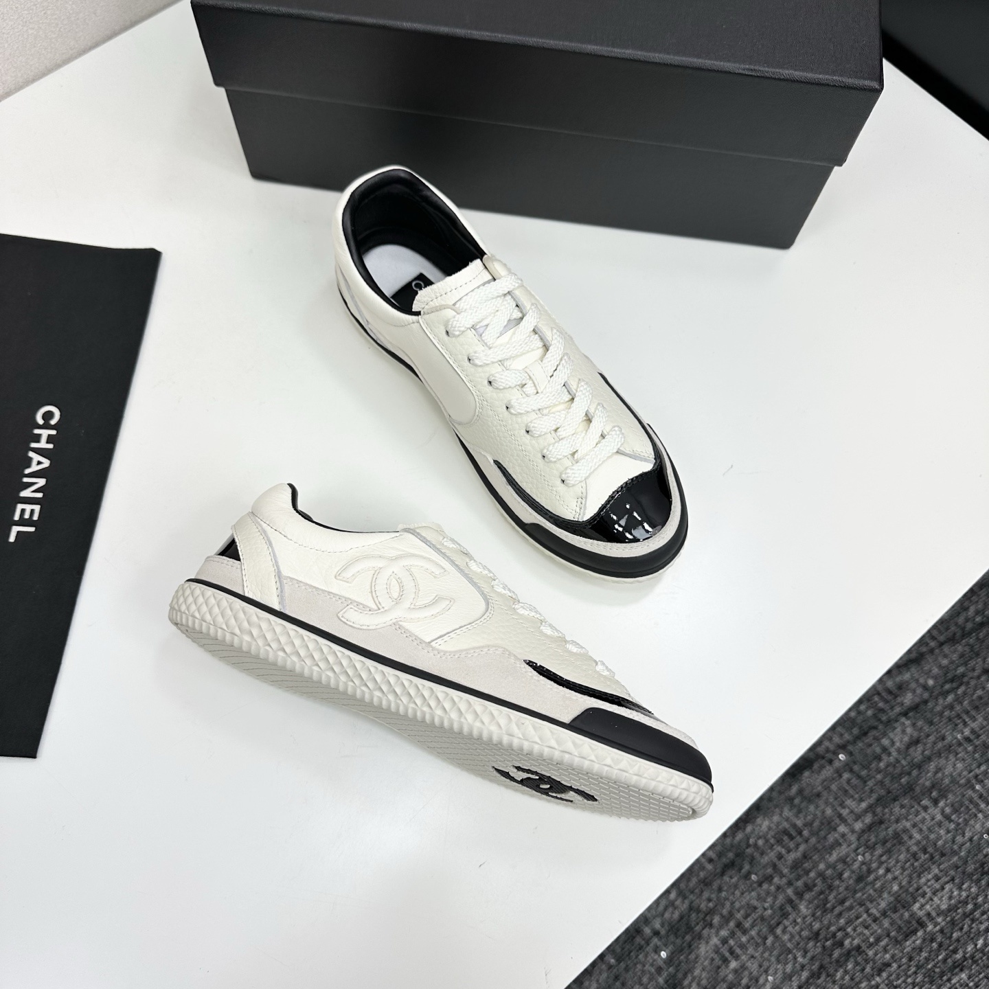 Pair of Chanel white leather sneakers with black patent toe caps and interlocking CC logo on a black display box - Hình 9