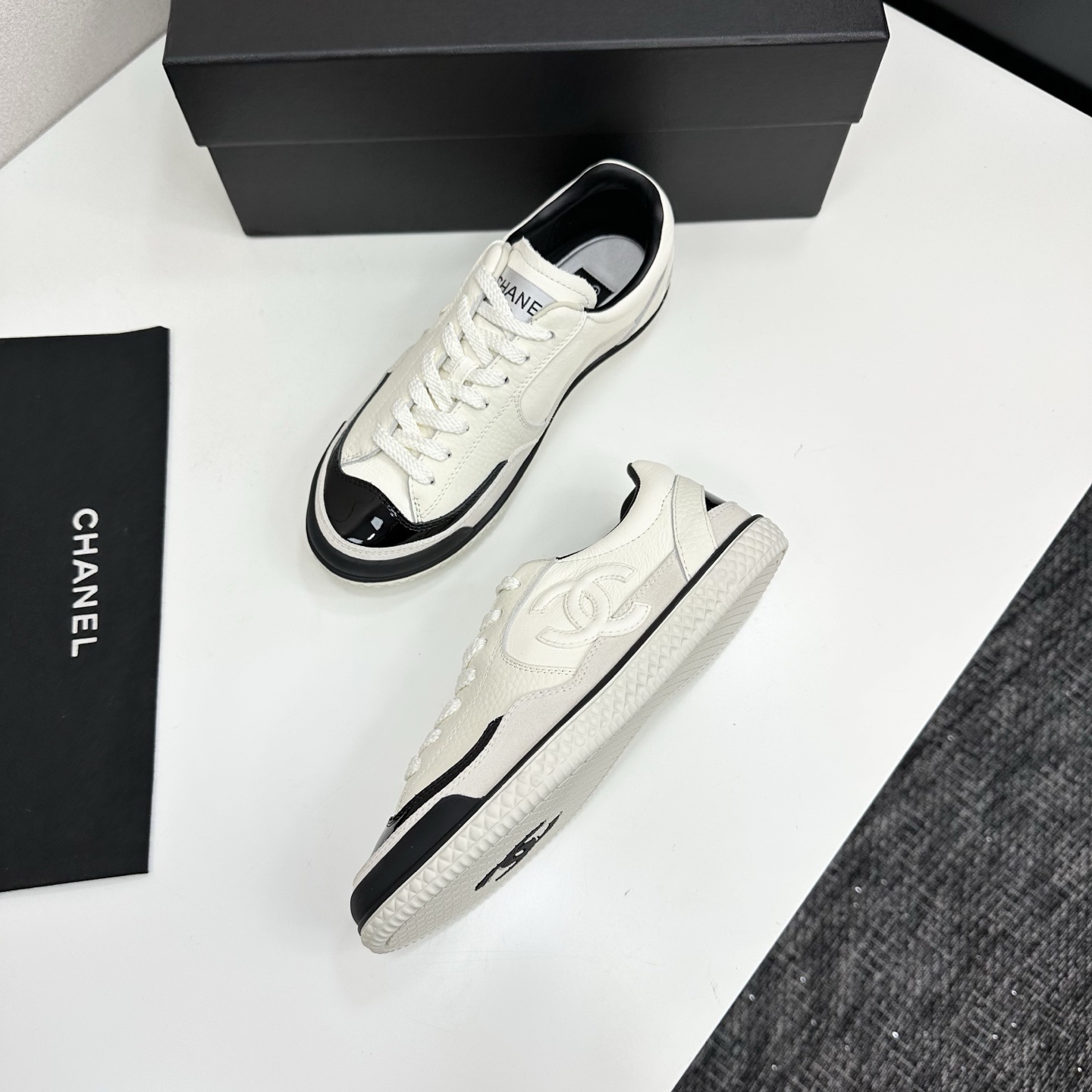 Pair of Chanel white leather sneakers with black patent toe caps and interlocking CC logo on a black display box - Hình 6