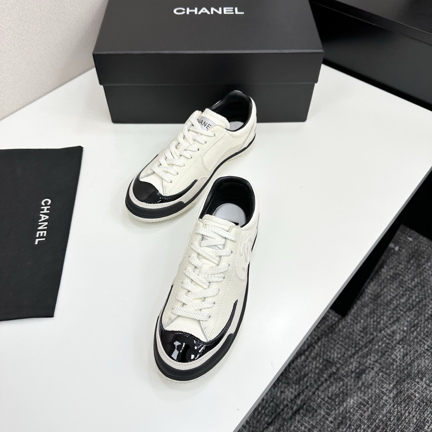 Pair of Chanel white leather sneakers with black patent toe caps and interlocking CC logo on a black display box - Hình 5