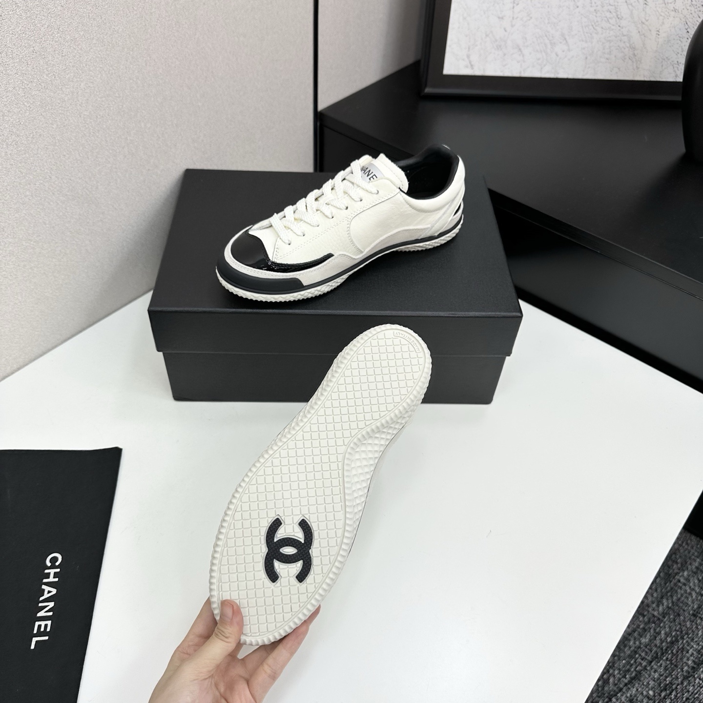 Pair of Chanel white leather sneakers with black patent toe caps and interlocking CC logo on a black display box - Hình 4