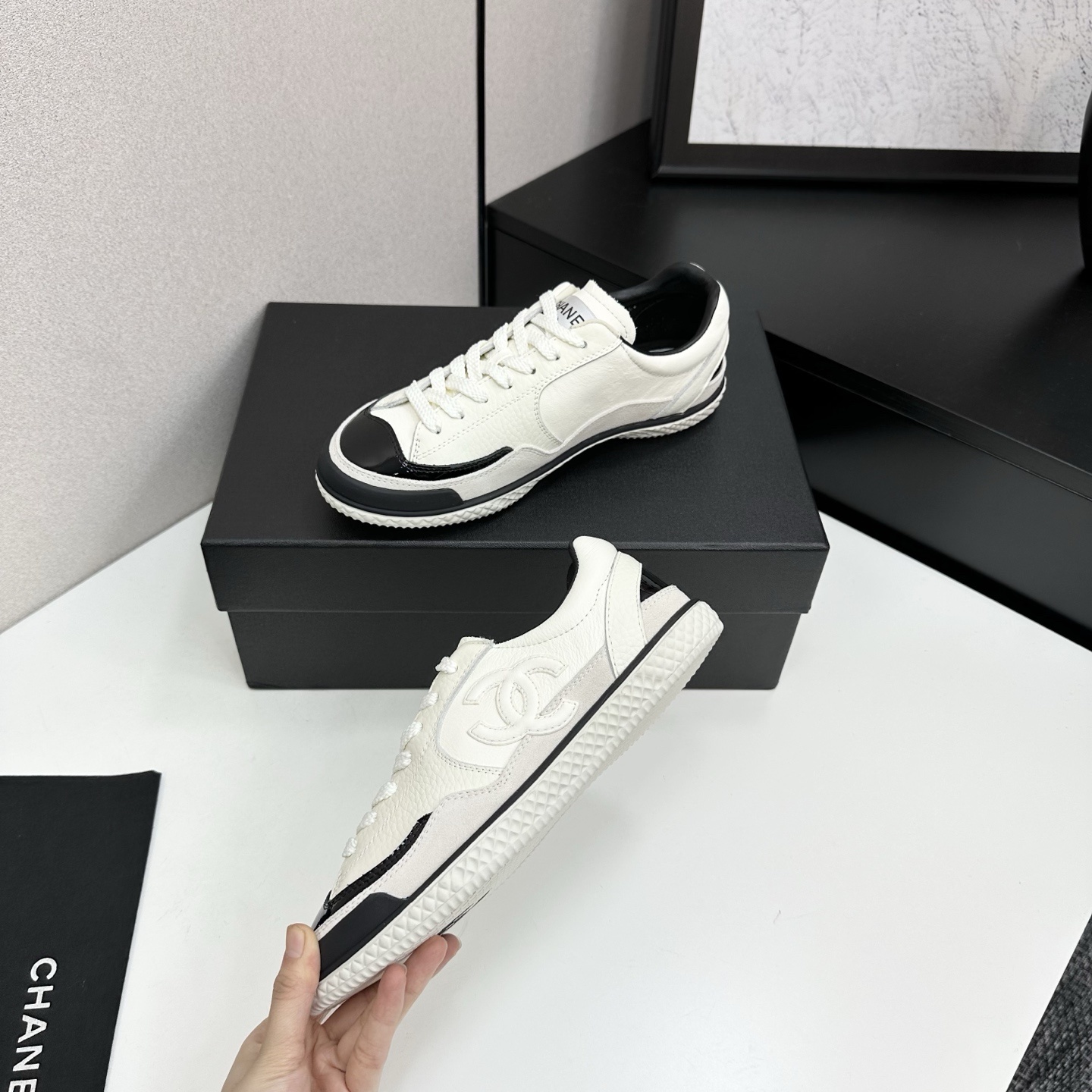 Pair of Chanel white leather sneakers with black patent toe caps and interlocking CC logo on a black display box - Hình 3