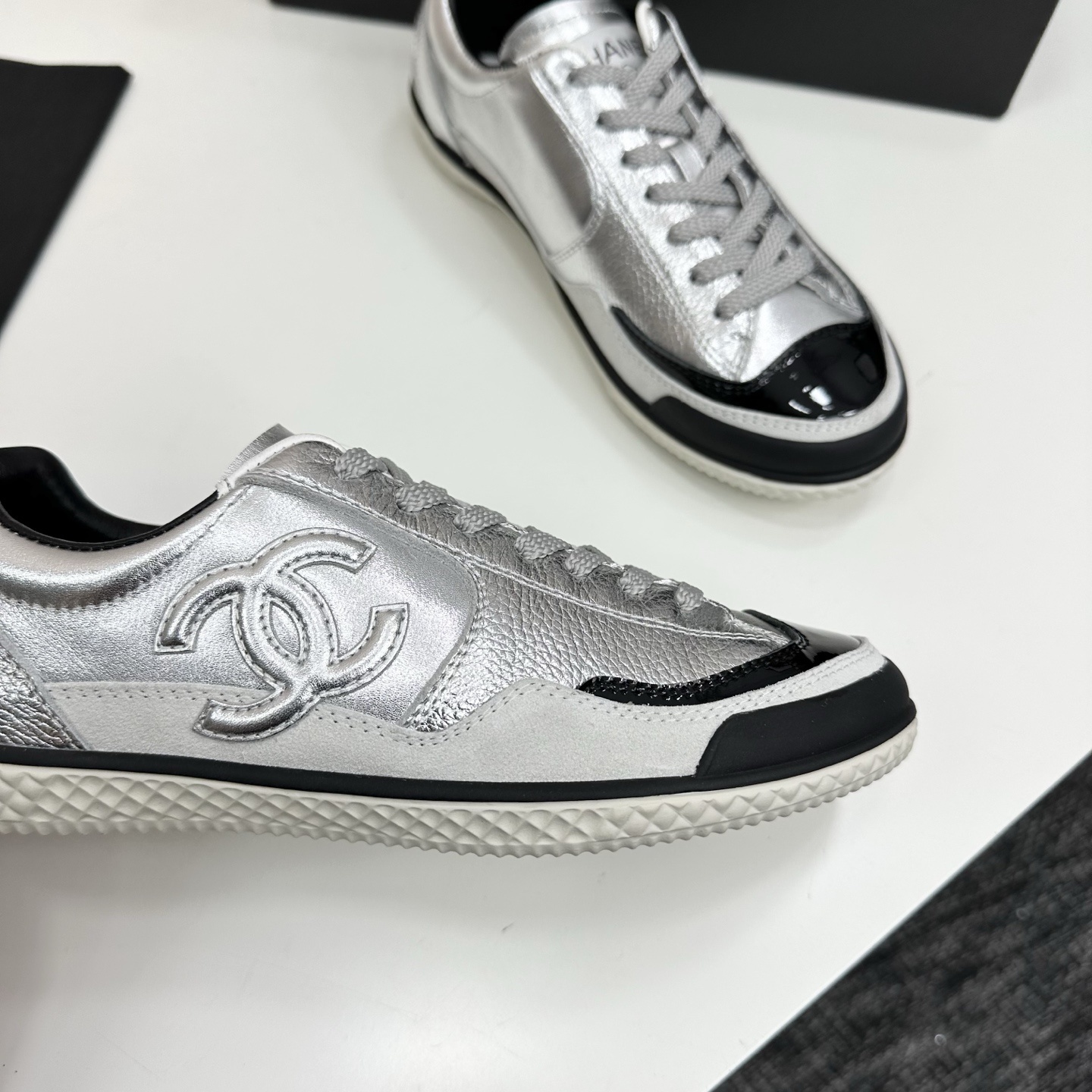 A pair of Chanel silver metallic sneakers with black accents and white soles displayed on a black box. - Hình 9