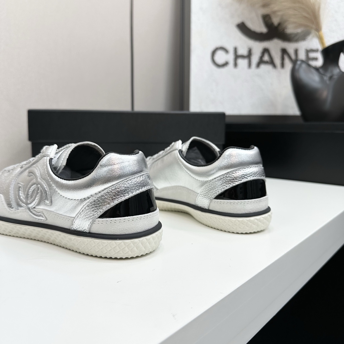 A pair of Chanel silver metallic sneakers with black accents and white soles displayed on a black box. - Hình 7