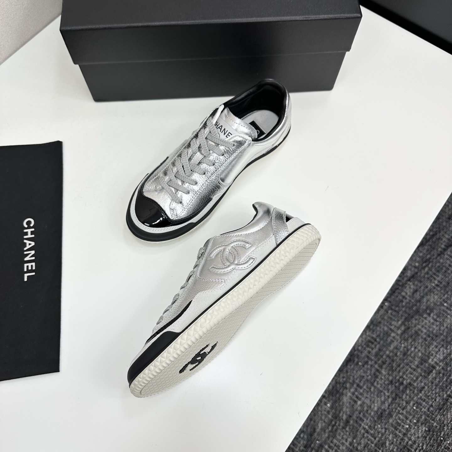 A pair of Chanel silver metallic sneakers with black accents and white soles displayed on a black box. - Hình 6