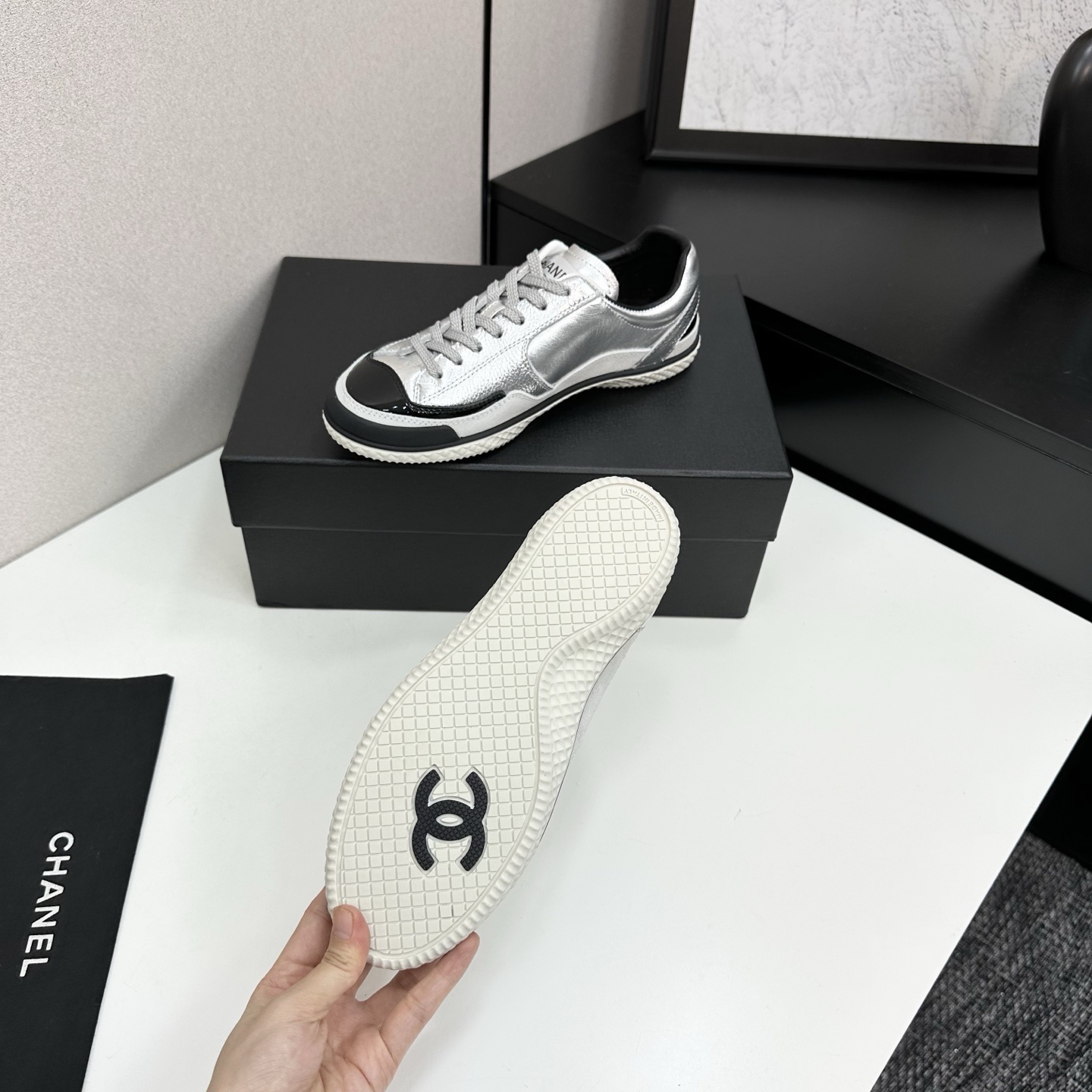 A pair of Chanel silver metallic sneakers with black accents and white soles displayed on a black box. - Hình 4