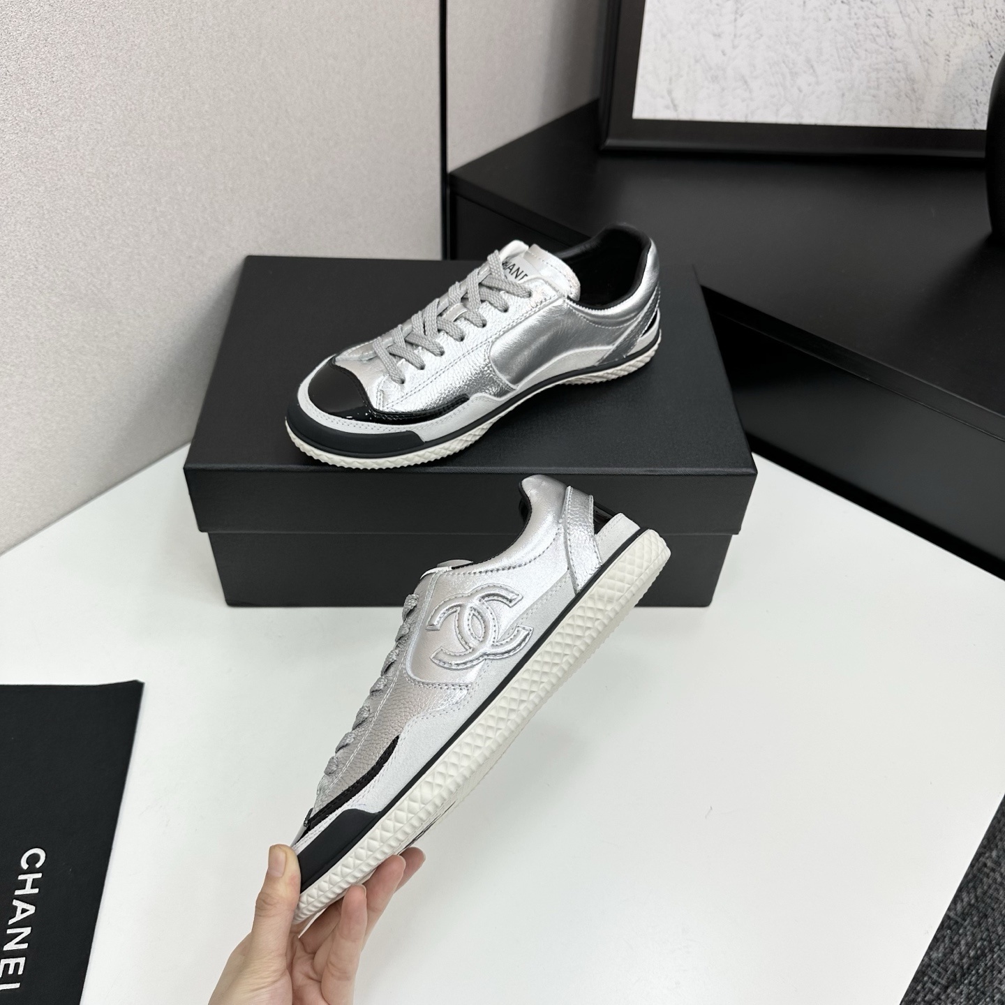 A pair of Chanel silver metallic sneakers with black accents and white soles displayed on a black box. - Hình 3