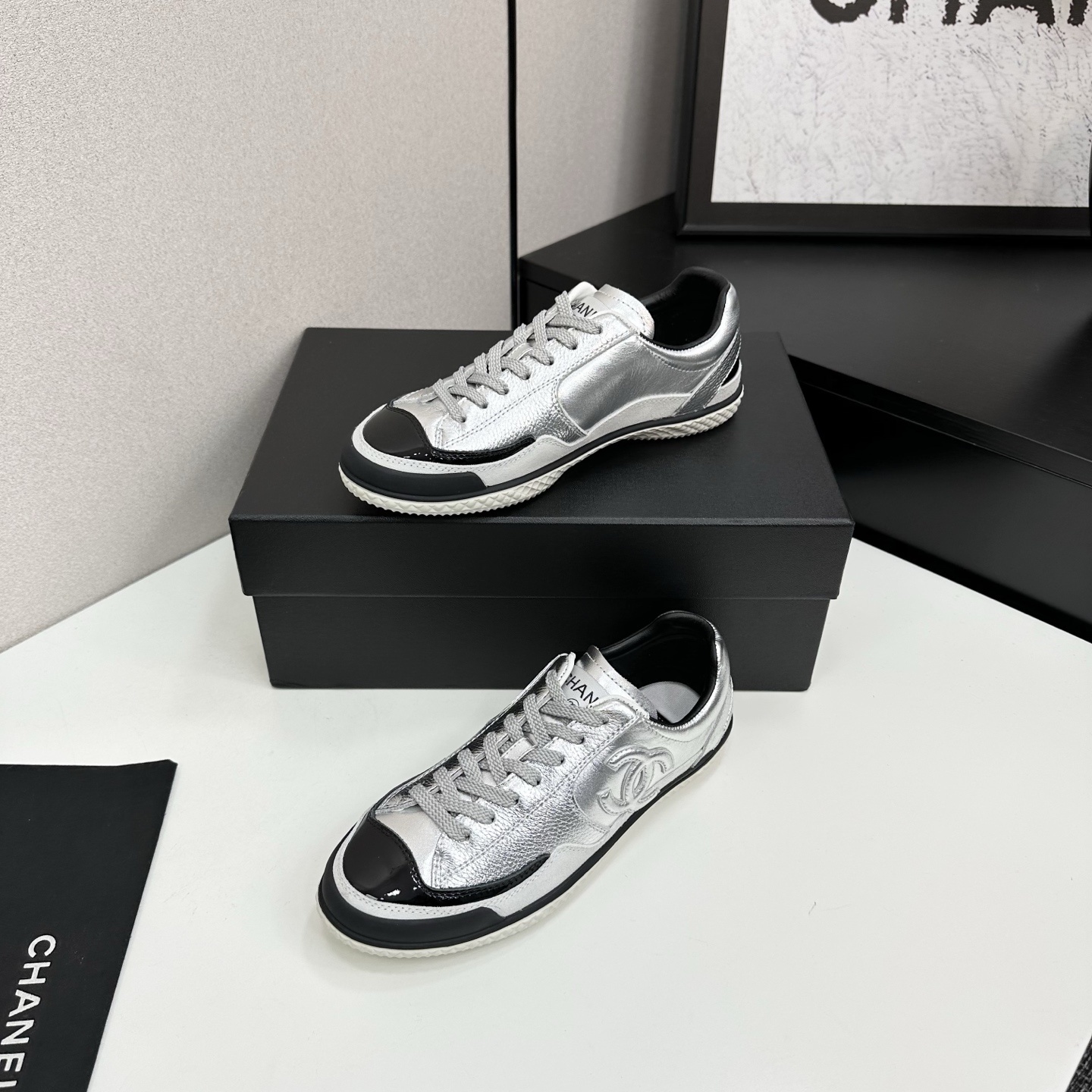 A pair of Chanel silver metallic sneakers with black accents and white soles displayed on a black box. - Hình 2