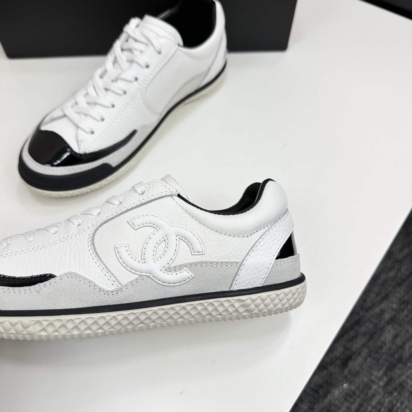Side view of white Chanel calfskin sneakers with black patent leather details and white interlocking CC logo on a black box. - Hình 7